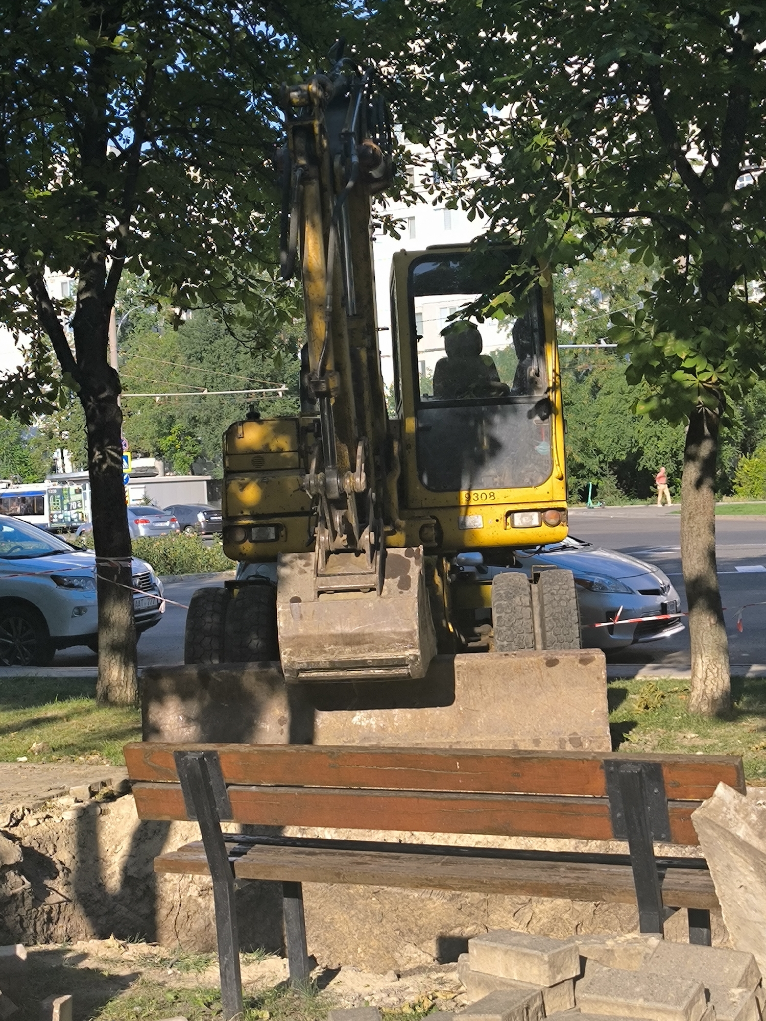 The image shows a yellow excavator parked on a street in Chișinău, Moldova. The excavator is positioned in front of a light gray sedan that appears to be partially under its bucket. In the background, there is a street with traffic, including a tram and several cars. Trees line the street, and buildings are visible in the distance. A wooden park bench is situated in the foreground, partially obscuring a dug-up area of ground. Two people are visible inside the excavator's cab. The scene appears to be a construction or road work area.