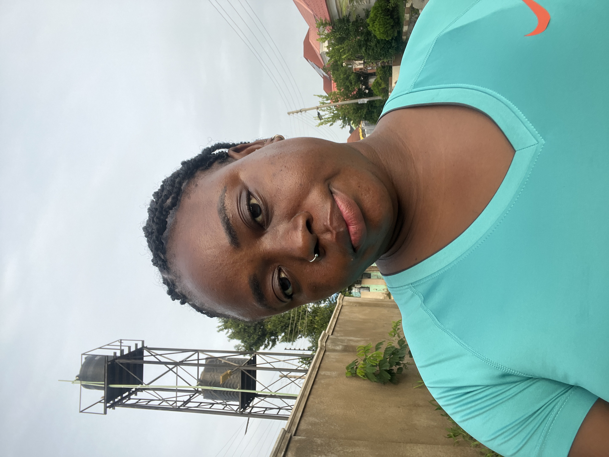 A medium shot photograph taken from the chest up shows a Black woman with braided hair and a nose ring posing for a selfie. She is wearing a turquoise v-neck t-shirt with a Nike logo on the right side of her chest. The background is an outdoor scene with a beige wall on the left, a tall metal water tower with two large black tanks, and residential buildings with red roofs on the right. The sky is overcast. The image appears to have been taken in Abuja, Nigeria.