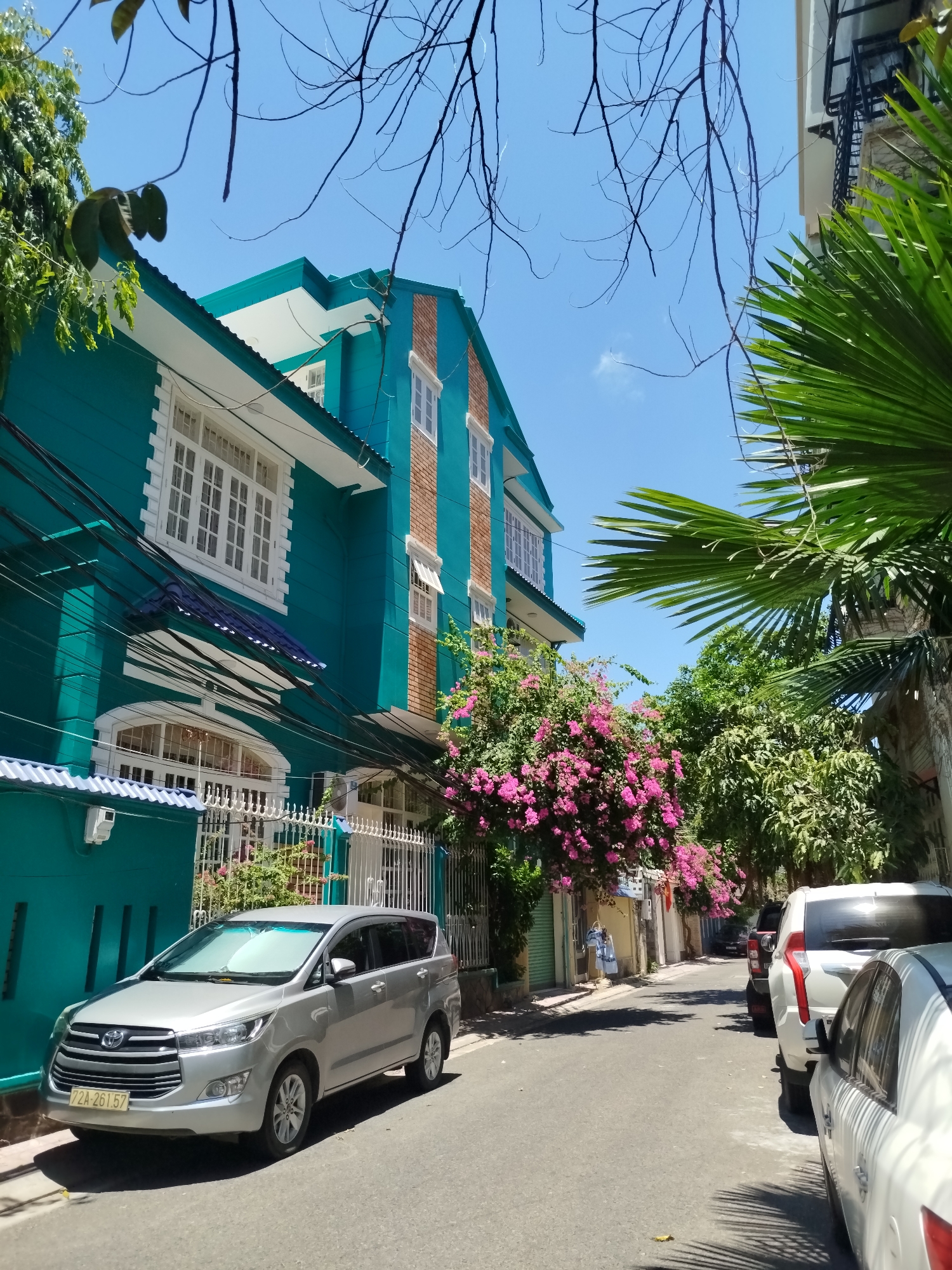 The image depicts a street scene in Ho Chi Minh City, Vietnam, characterized by a teal-colored building on the left with white trim and brick accents on an upper section. The building has a prominent fence along the front. A silver Toyota Innova is parked in front of the building, facing the camera. The license plate appears to read "72A-261.57". The street itself is narrow, with other vehicles parked along the right side. Lush greenery, including palm trees and flowering bushes with pink blossoms, lines the street. Overhead, utility wires crisscross. The sky is clear and blue. There are also other buildings along the street, although these buildings are not visible in great detail due to the distance and some obstruction by vegetation.