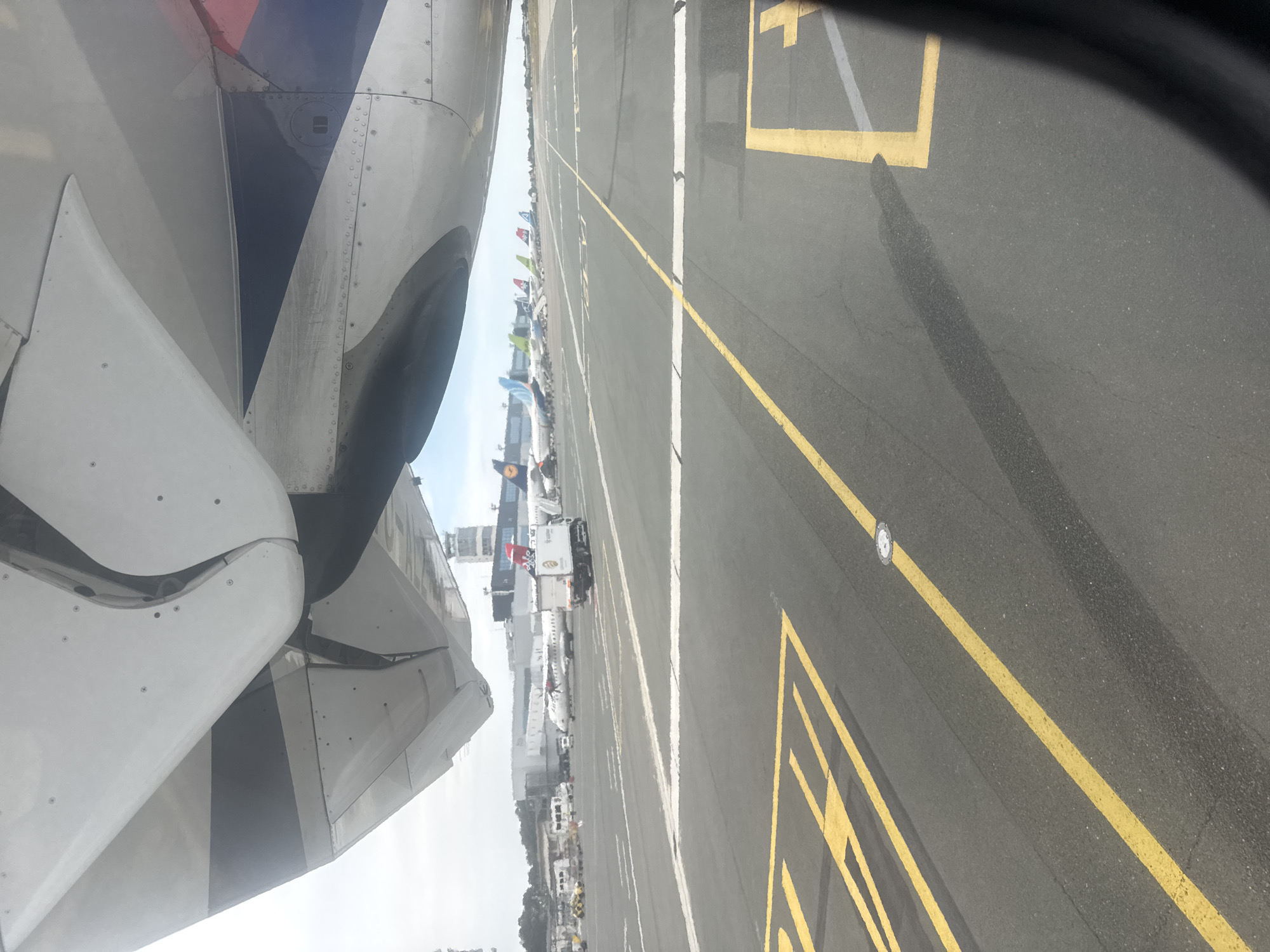This image shows a view from an airplane window, looking out at the tarmac of an airport. The left side of the frame is dominated by a large section of the airplane's fuselage and wing, a portion of which is in shadow. The wing is white and appears to be made of metal panels. The fuselage shows sections of blue and red paint.
The right side of the image displays the airport tarmac, marked with yellow and white lines. In the distance, several other airplanes are visible, parked at gates or taxiing. Some are small, while others are larger commercial jets. A few service vehicles and jet bridges are also present. The sky is bright, suggesting it is daytime, and the weather appears to be clear. The location is identified as Surcin Urban Municipality, Serbia, likely Belgrade Nikola Tesla Airport. The scene depicts the typical environment of an airport apron during operating hours.