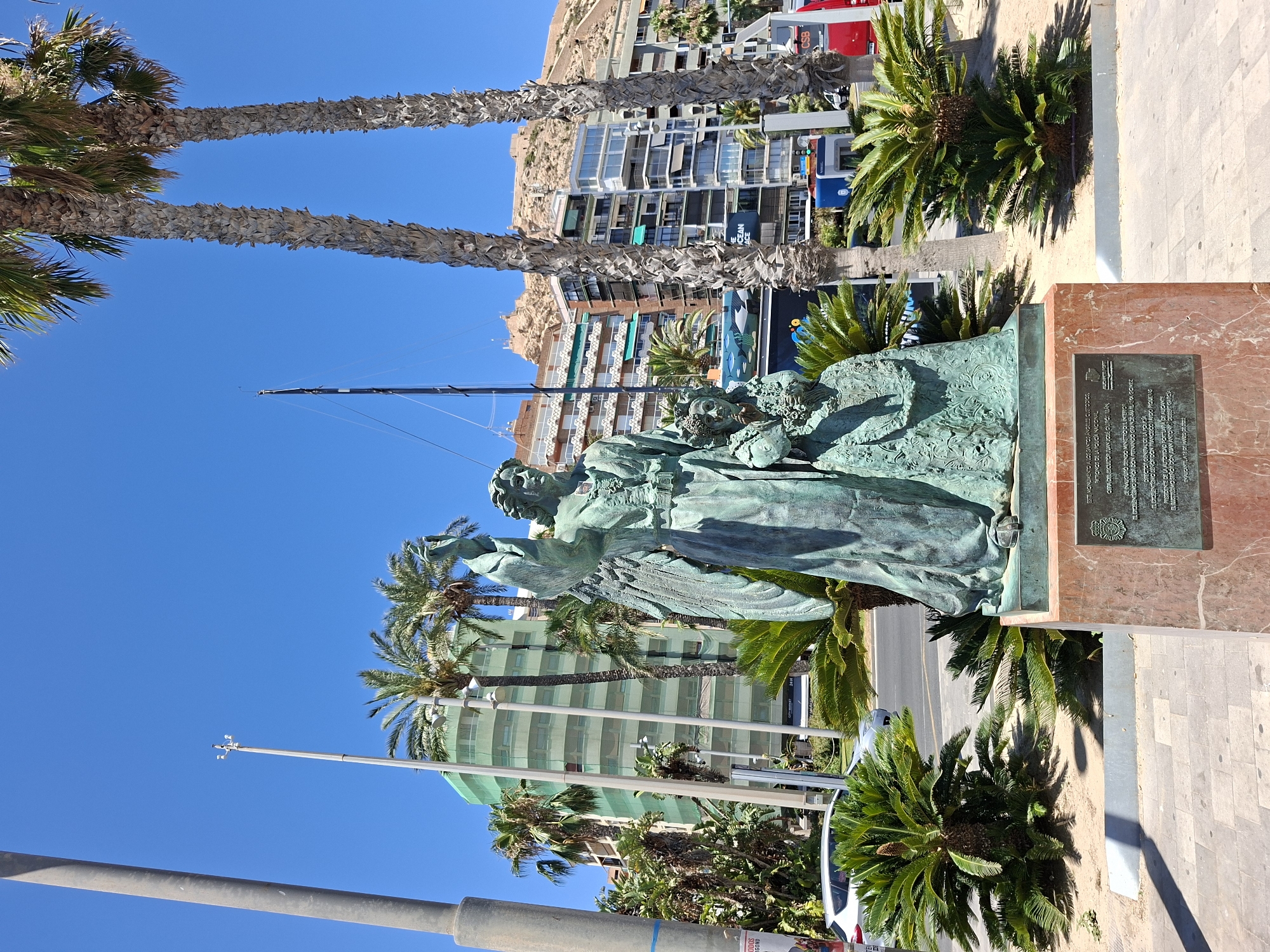 The image shows a bronze statue of an angel with outstretched wings and a crown, standing next to a young girl. The statue is situated on a marble pedestal with an inscription plaque. The statue is located in front of a street with palm trees and a clear blue sky. In the background, there are multi-story buildings, some of which are covered with green netting, suggesting ongoing construction or renovation. Further in the distance, a castle or fortress is visible on a hilltop. This monument is located in Alicante, Spain, specifically at the Explanada de España. The inscription on the plaque states that the monument was inaugurated on January 16, 2023. The environment appears to be a sunny day in a coastal city, with cars and a red bus visible in the background.