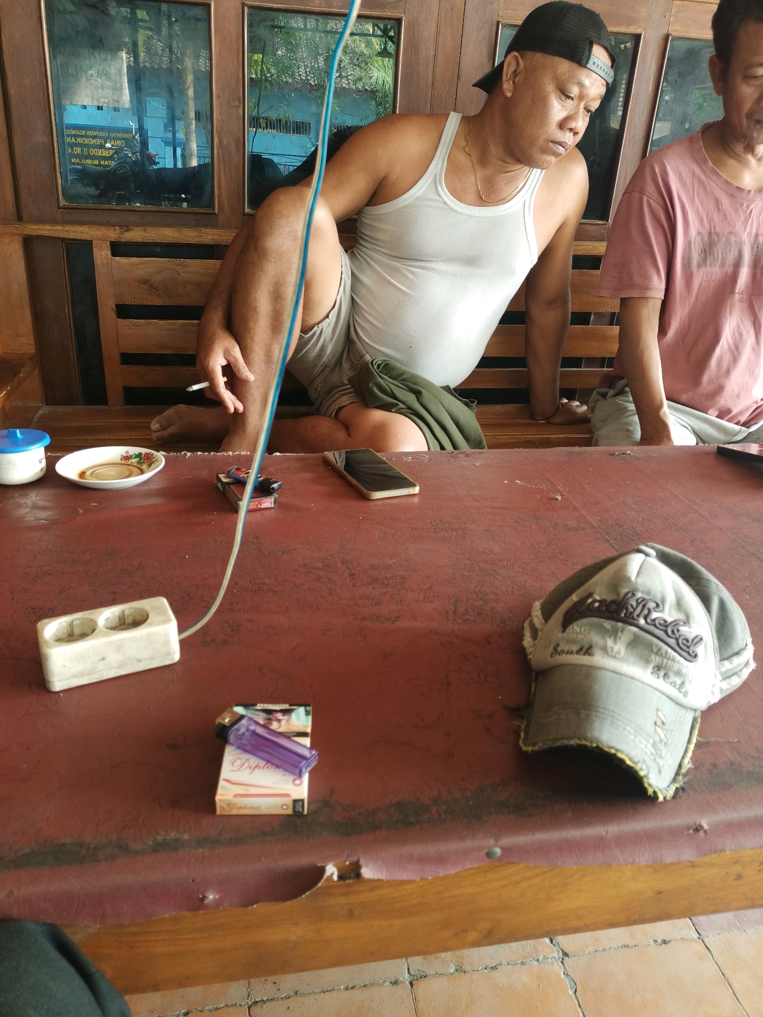 Two men are seated at a red table, likely outdoors, with a wooden structure behind them. The man on the left wears a white tank top, grey shorts, and a black baseball cap, and is holding a cigarette. The man on the right wears a pink t-shirt. On the table are various items including a pack of cigarettes with a purple lighter, a small white bowl with liquid, a white object with two electrical outlets, a smartphone, and a baseball cap. The background reveals wooden benches and glass panels reflecting trees and buildings, suggesting an open-air setting. The environment appears to be a public space or a communal area in Sumber Bendo, Indonesia.