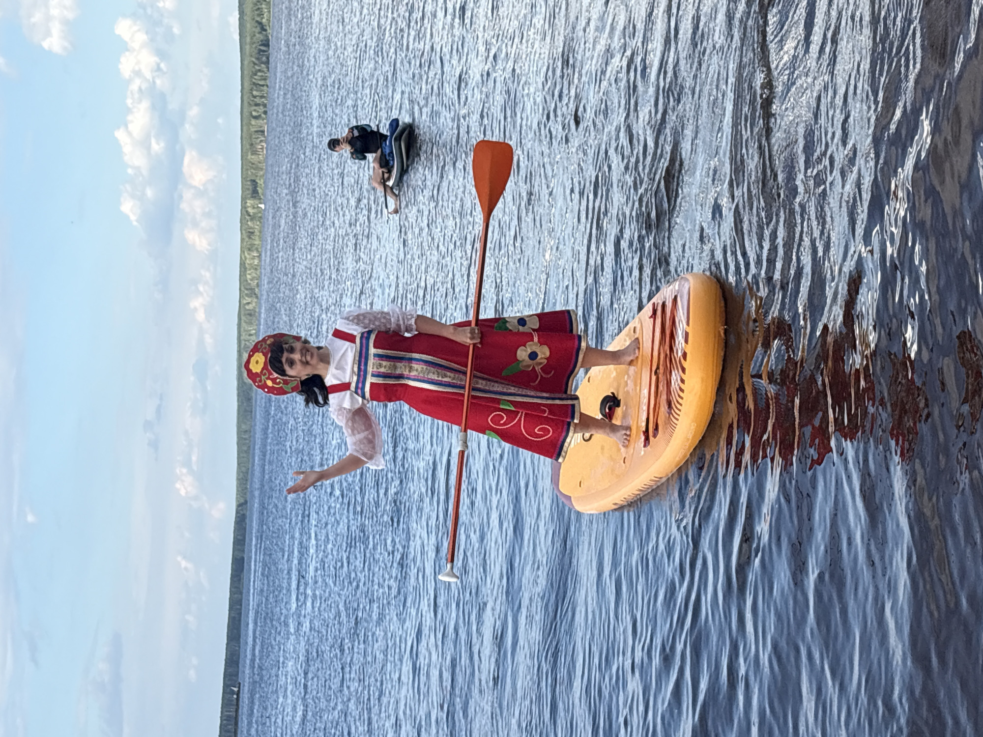 In this photograph, two people are paddleboarding on a calm lake in Chaikovsky, Russia. The foreground features a woman in a traditional red and white embroidered dress and a red, floral-patterned headdress, standing on an orange paddleboard. She holds an orange paddle, its tip submerged in the water, and is gesturing with her left hand. In the background, another person is seen paddleboarding on a black board. The lake reflects the sky, which is partly cloudy with white cumulus clouds against a pale blue backdrop. The shoreline in the distance is lined with a dense green forest. The water is a deep blue, with visible ripples and reflections. The image appears to be taken during the daytime, under clear weather conditions. The woman's attire suggests a cultural or celebratory context, and her pose conveys a sense of graceful engagement with the activity.