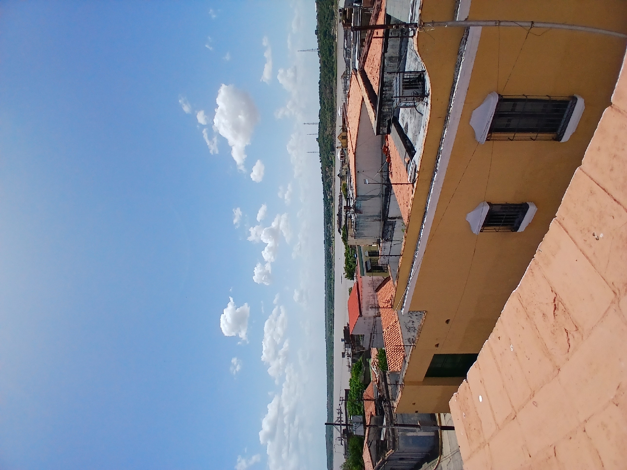 A slightly skewed, eye-level outdoor shot shows a cityscape, possibly in Ciudad Bolivar, Venezuela, marked by various buildings with orange-tiled roofs and yellow walls juxtaposed against a blue sky with scattered white clouds. In the distance, there is a treeline and some communication towers. The building on the right has two barred windows. There are no visible people or explicit actions. The interaction between buildings and sky constitutes the primary visual element.