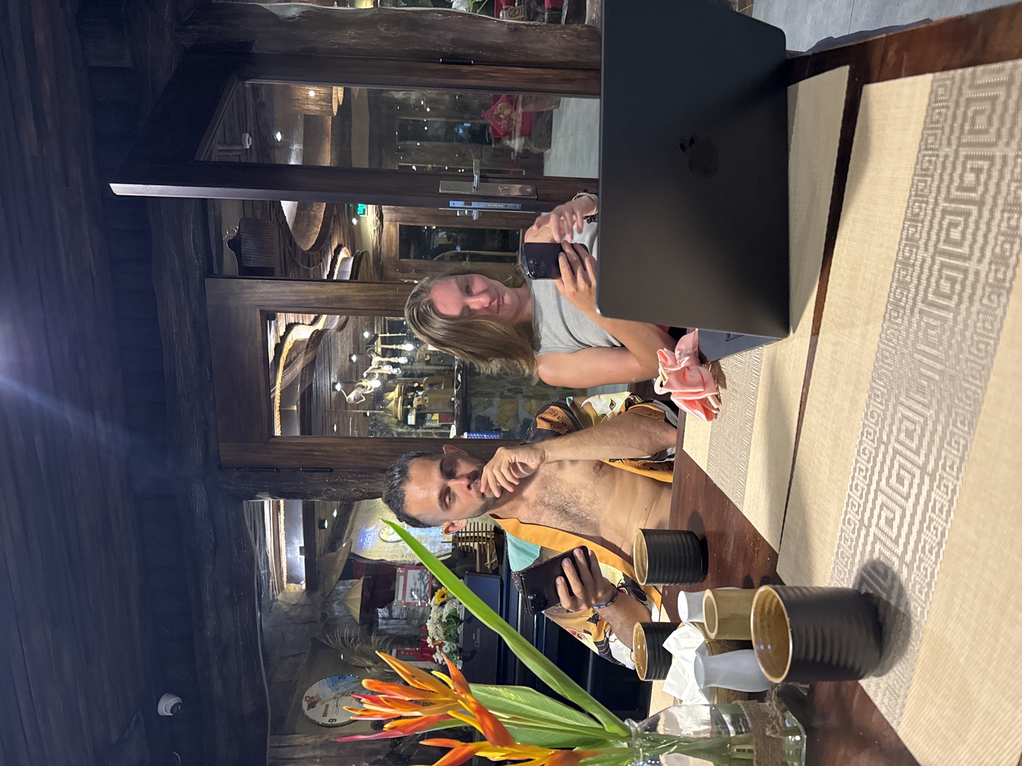 An exterior shot captures a scene with a man and a woman sitting at a table in what appears to be an outdoor seating area of a restaurant. The man, shirtless and wearing a patterned robe, is engrossed in his smartphone, holding it with his left hand. Beside him, a woman in a gray tank top is also looking at her smartphone. In front of them, a laptop is open on the table, its screen obscured. The table is set with placemats, cups, and a vase of vibrant orange and yellow flowers. The background reveals a rustic, wooden interior with exposed beams and traditional Vietnamese architectural elements, suggesting a location in Nam Hoa Lư Ward, Vietnam, possibly a restaurant or a guesthouse.