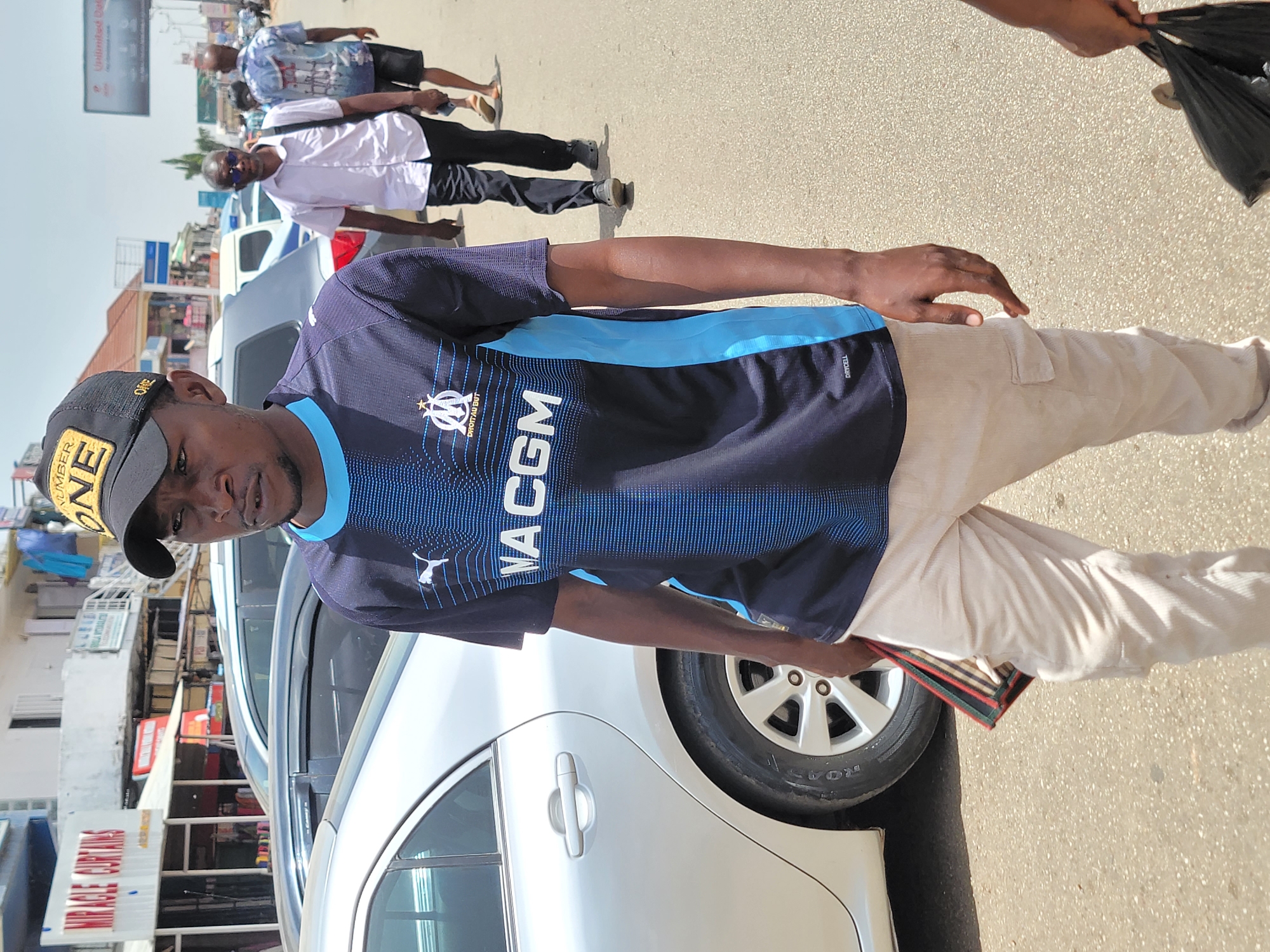 A man stands in front of a silver sedan on a street in Jos, Nigeria. He wears a dark blue soccer jersey, light-colored pants, and a black baseball cap. He holds what appears to be a wallet or small bag. Several people walk in the background. Buildings are visible across the street.