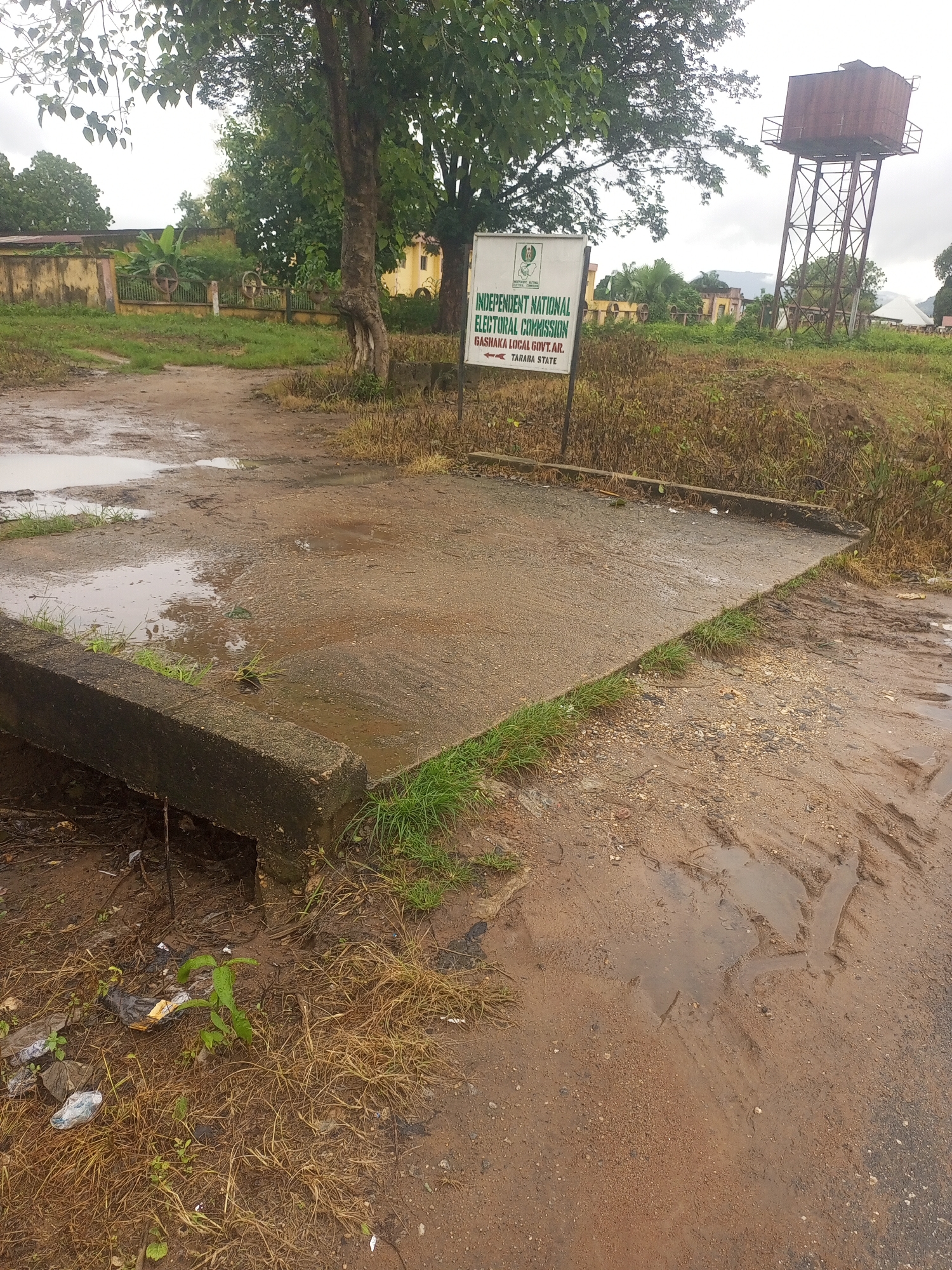 The image depicts an outdoor scene in Serti, Nigeria. A sign in the foreground reads "INDEPENDENT NATIONAL ELECTORAL COMMISSION GASHAKA LOCAL GOVT.AR. TARABA STATE" with an emblem and an arrow pointing to the left. The ground is uneven and muddy, with puddles of water indicating recent rain. A concrete structure, possibly a bridge or drainage, runs along the left side of the frame. Vegetation, including grass and trees, is present throughout the scene. In the background, there are buildings and a tall, rusty water tower. The overall environment appears somewhat neglected and weathered.