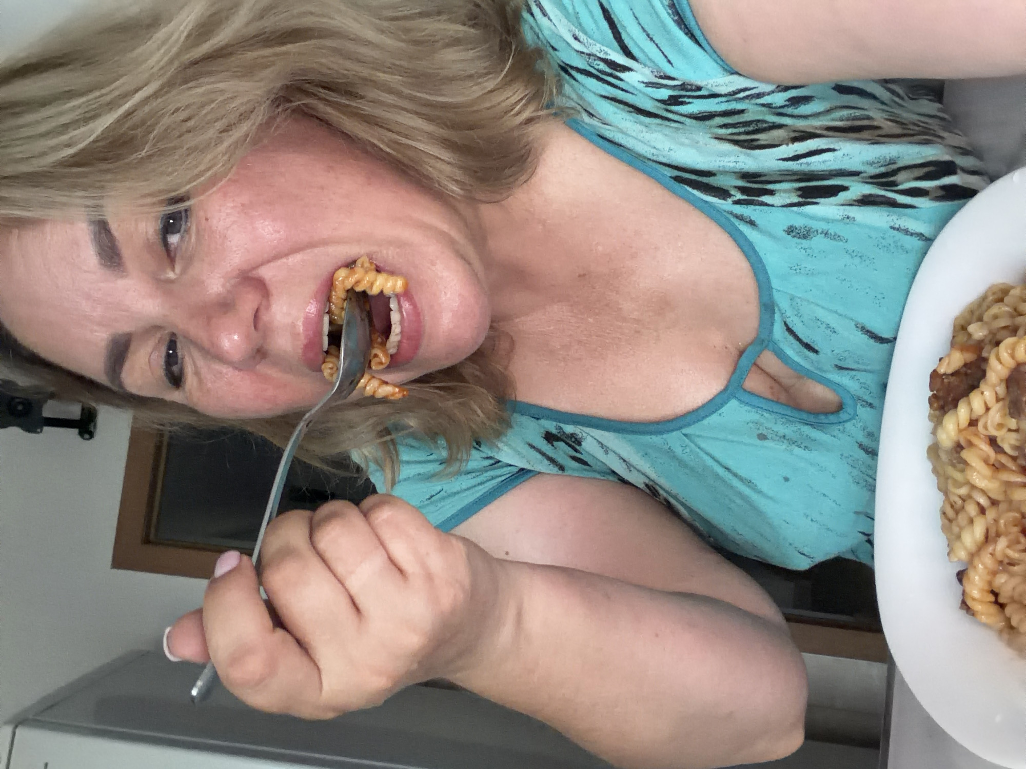 A middle-aged woman with blonde hair is captured in a close-up selfie as she eats pasta from a spoon. She is wearing a blue and black patterned top with a keyhole neckline. The background shows a glimpse of a kitchen or dining area, with a refrigerator on the left and a doorway to the right. The woman is leaning forward, bringing a spoonful of pasta towards her mouth. The lighting is bright, suggesting an indoor setting during the daytime. The location is Leningradskaya, Russia.