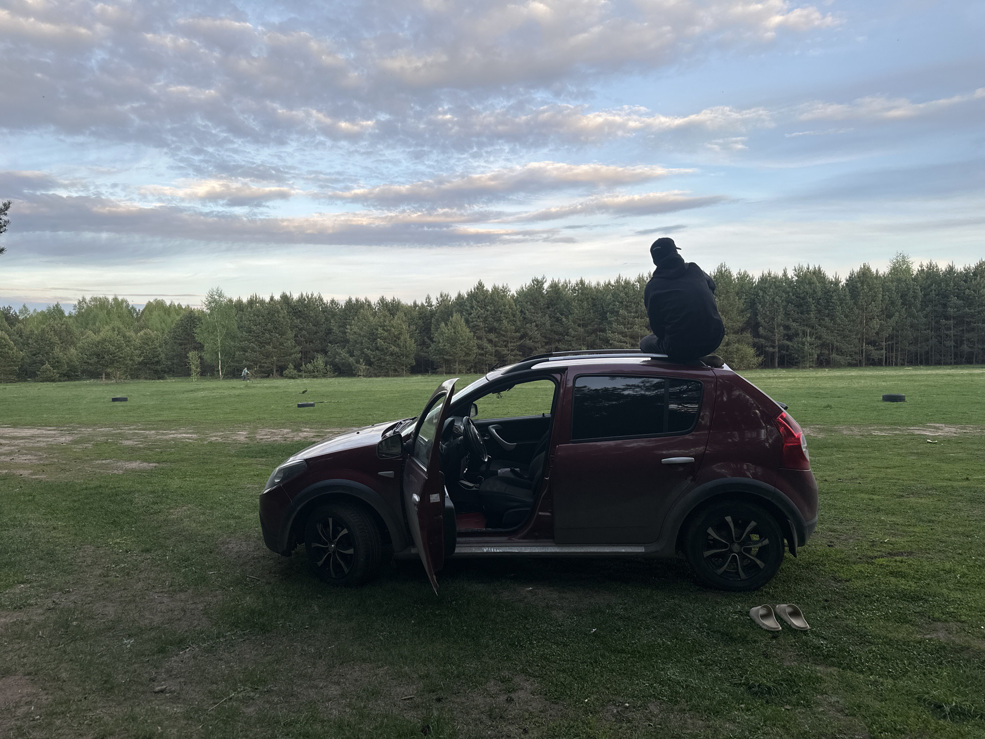 The image shows a red Dacia Sandero Stepway parked on a grassy field in a natural area, possibly near Chaikovsky, Russia. A person wearing dark clothing and a cap is sitting on the roof of the car, facing away from the camera. The driver's side door of the car is open. In the background, there is a line of trees, a field with some objects that appear to be tires, and a cloudy sky. A pair of sandals are on the grass near the rear of the car. The overall impression is one of a relaxed outdoor setting.