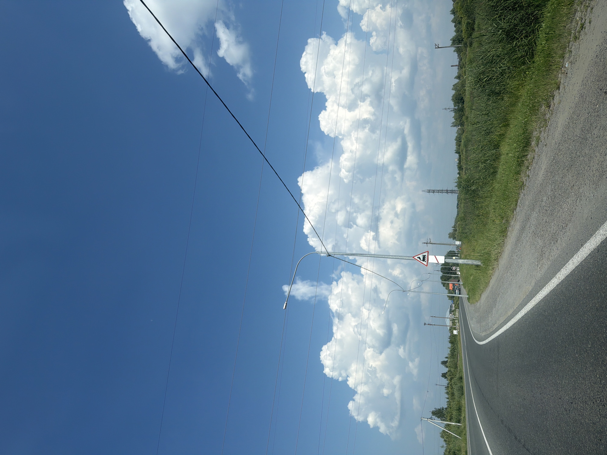 The image depicts a bright, sunny day on a rural road near Tobolsk, Russia. The sky is a vibrant blue, dotted with large, fluffy white clouds. Power lines stretch across the upper portion of the frame. A paved road curves to the left, with a gravel shoulder on the right. Along the side of the road, tall green grass and reeds grow abundantly. In the distance, trees form a dense line. Several utility poles and a street lamp are visible. A triangular road sign with an image of a train is mounted on a pole. In the far distance, industrial structures like cooling towers or radio masts can be seen. There are no people or vehicles clearly visible in the immediate foreground. The overall scene suggests an open, natural landscape with signs of infrastructure.