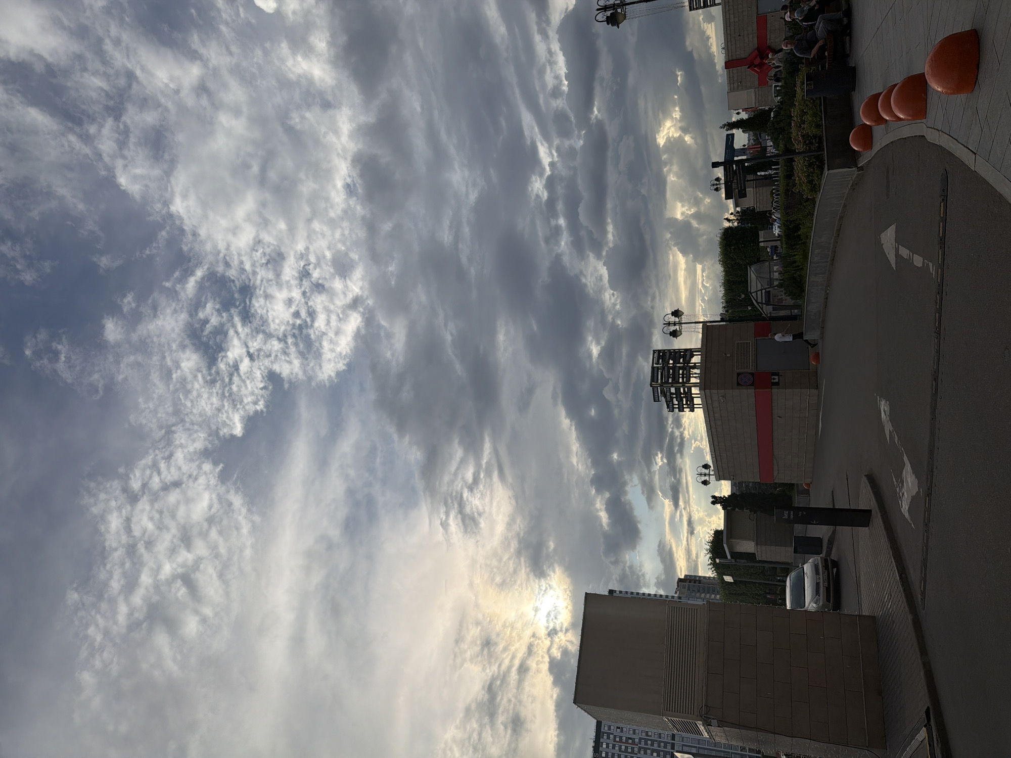 The image shows an outdoor scene with a sky full of dramatic clouds, suggesting either an approaching storm or the aftermath of one, with sunlight breaking through in some areas. In the foreground, there is a paved area with directional arrows painted on the ground, indicating traffic flow. To the left, a large, modern building facade made of textured concrete blocks is visible. A light-colored car is parked on the left side of the road. Further back, several other buildings and structures are present, including a building with red trim and a large red ribbon decoration, possibly indicating a sale or event. There are streetlights and some greenery. On the right side of the image, a paved walkway area has several large, orange, spherical bollards. A group of people are seated on a bench in this area, with one man visible from the chest up. Another person is walking away from the camera on the left side. The overall impression is a public outdoor space in an urban environment, possibly a shopping or entertainment complex, under a moody sky.