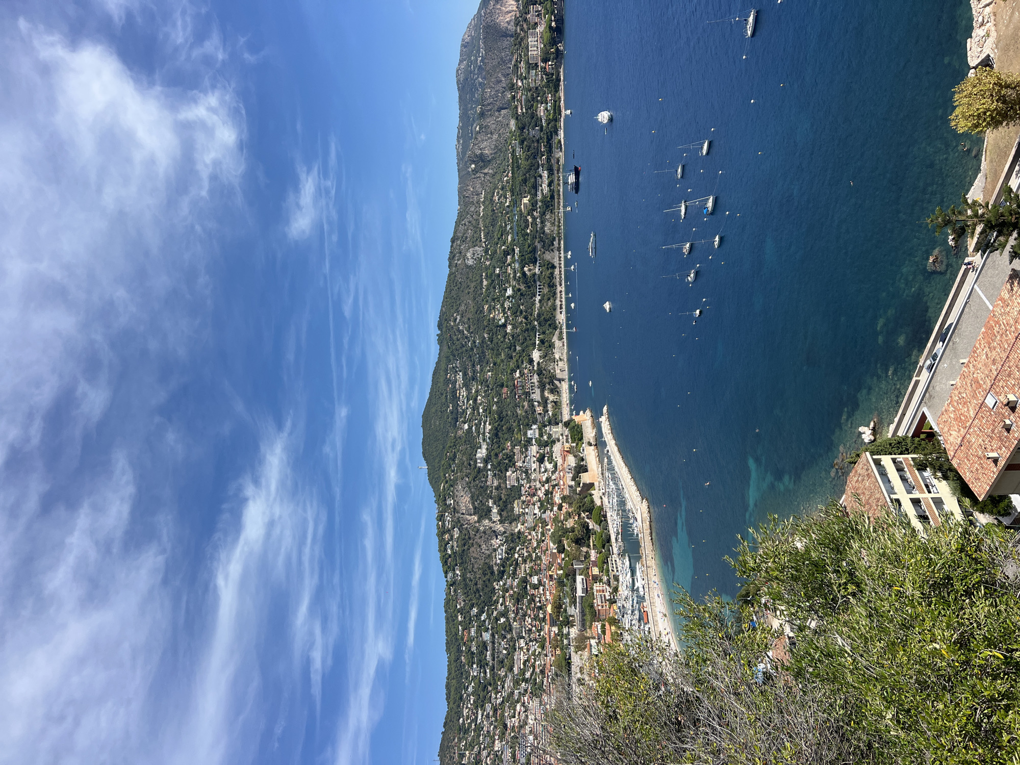 This is an aerial, high-angle, eye-level shot of a coastal city in Nice, France, on a sunny day with a few wispy clouds in the sky. The scene shows a bay with deep blue water dotted with numerous boats, ranging from small sailboats to larger yachts. A prominent curved marina juts into the bay, filled with many docked sailboats and motorboats.
The city itself is built on a steep, green hillside that slopes down towards the water. Buildings of various sizes and colors, mostly with terracotta roofs, are densely packed along the slopes and closer to the waterfront. Some buildings appear to be residential, while others may be commercial or hotels. A road runs along the edge of the water in some areas, and a small beach with a few people is visible near the marina.
In the foreground, the edge of a lush green tree with bare branches extends into the frame from the left, partially obscuring the view of the city below. Another building with a yellow facade and terracotta roof is visible in the lower left corner of the image, closer to the viewer.
The overall impression is one of a picturesque Mediterranean coastal town, bustling with activity on the water and nestled against a verdant, hilly landscape.