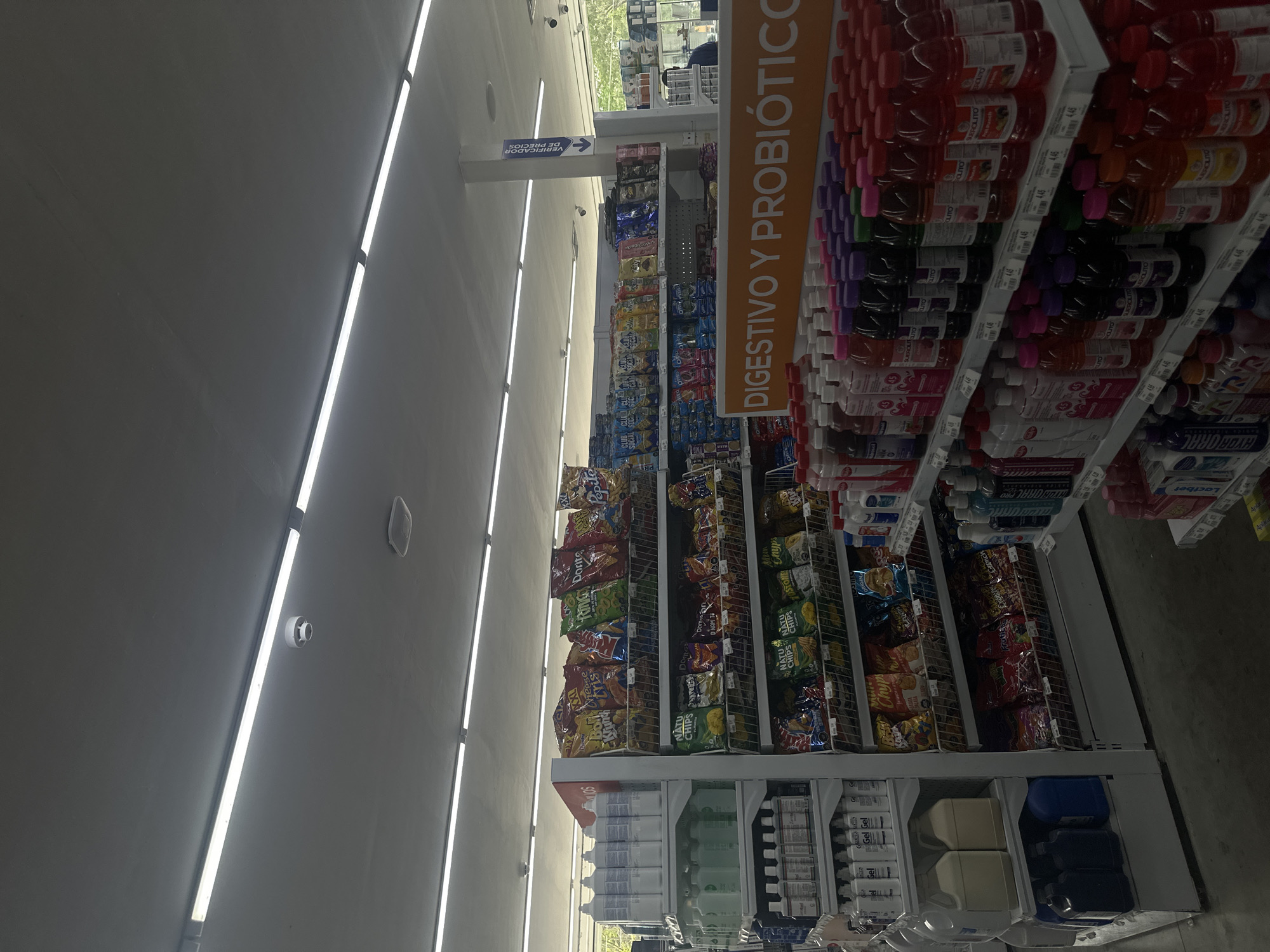 The image depicts the interior of a grocery store, presumably located in Parroquia Ocumare, Venezuela, given the presence of Spanish-language signage. The focus is on the aisles, which are lined with shelves stocked with a variety of snack foods and beverages.
Prominently featured is a sign advertising "DIGESTIVO Y PROBIÓTICO," suggesting a section dedicated to digestive aids and probiotic products, consisting of drinks. Adjacent to this, shelves are filled with various brands and flavors of chips, displayed in their packaging.
Below the chips, a lower section houses larger containers of liquids and other products, possibly cleaning supplies or larger format beverages. Fluorescent lighting fixtures are visible on the ceiling, providing illumination to the store. The scene conveys an everyday shopping environment within a typical grocery store setting.