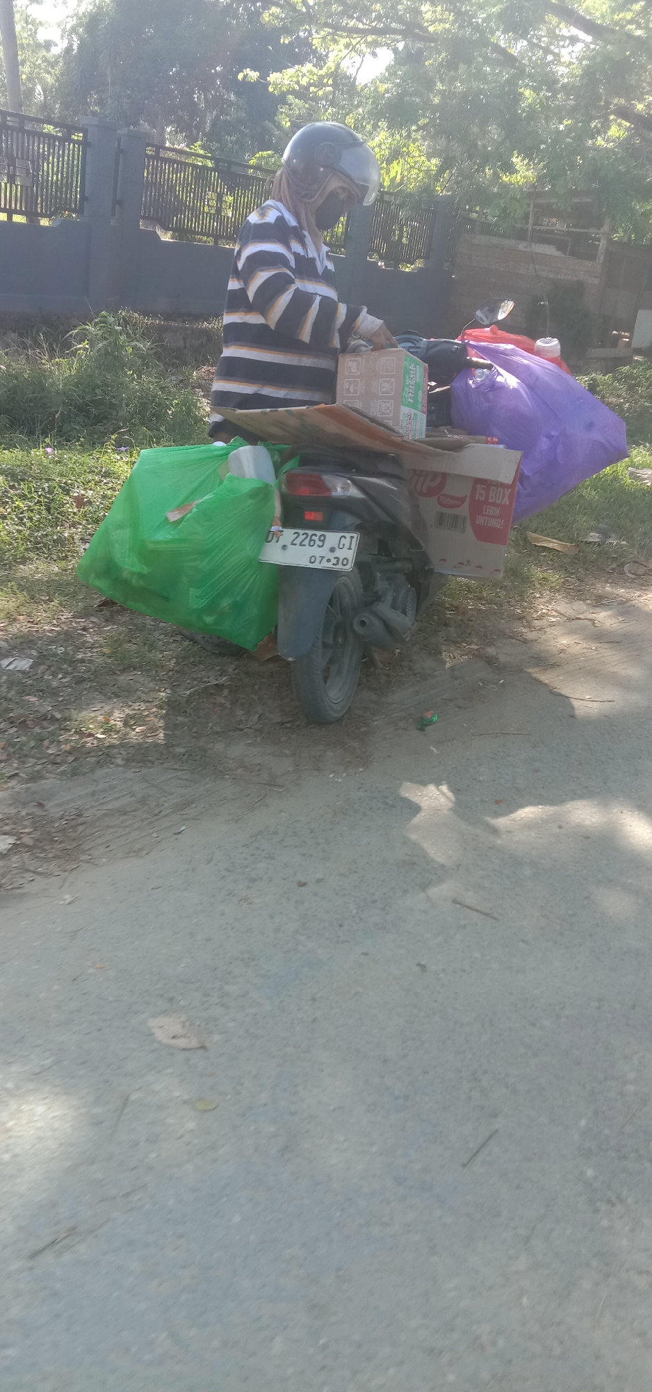 A person wearing a striped long-sleeved shirt, a black helmet, and a mask is standing next to a motorcycle parked on the side of a road in Kendari, Indonesia. The motorcycle is loaded with several large items, including a bright green plastic bag on the left, two cardboard boxes on the right, and a large purple plastic bag. The boxes appear to contain goods, with one box labeled "15 BOX LEBIH UNTUNG!". The person is reaching towards one of the boxes, possibly arranging or securing the cargo. The environment suggests an outdoor setting with vegetation and a fence in the background, under bright sunlight, casting shadows on the paved road. The license plate on the motorcycle reads "DT 2269 GI 07-30".