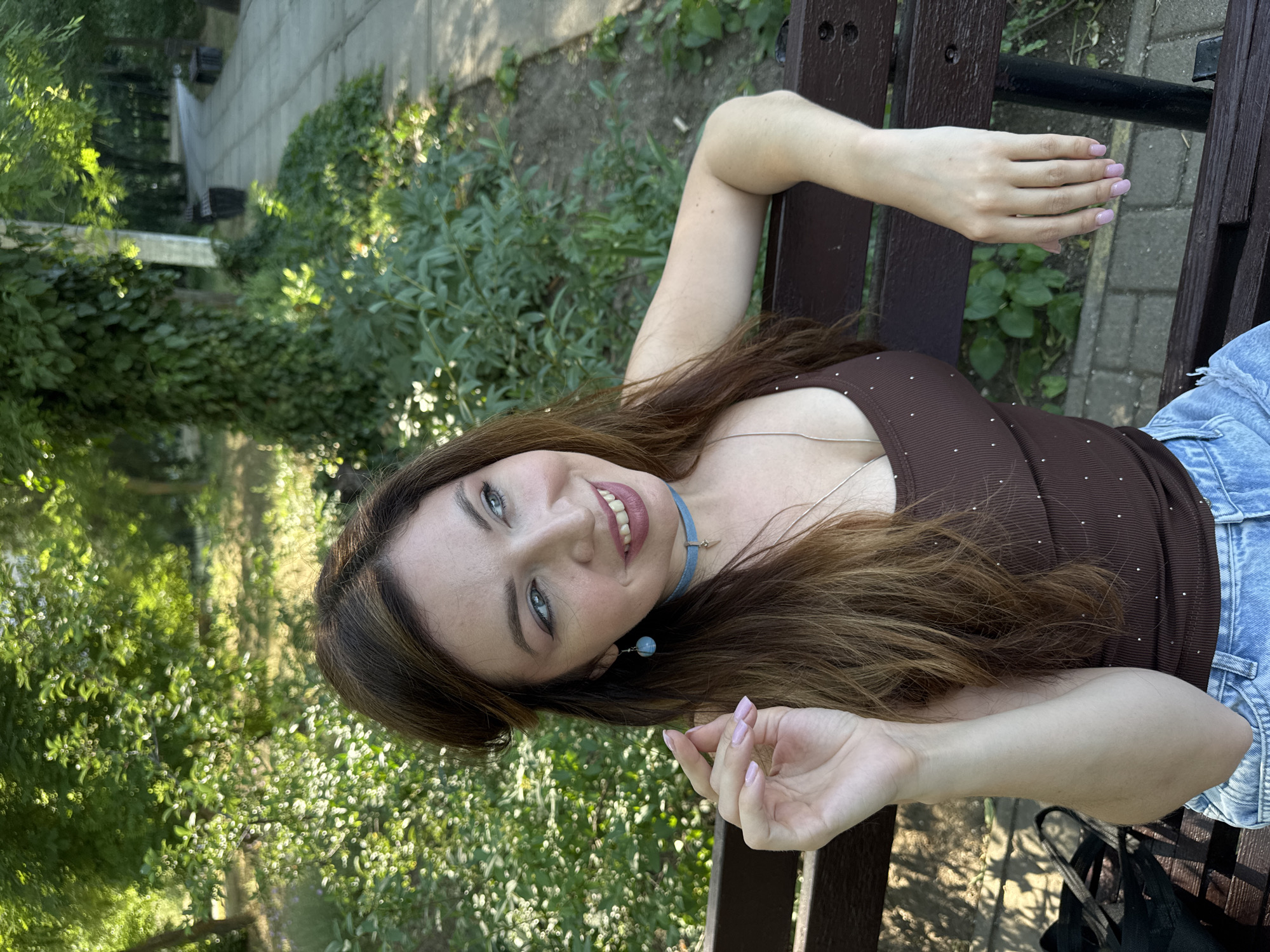 A young woman is lying on a wooden bench outdoors, with her head towards the left of the frame and her body extending to the right. She has long, dark brown hair, blue eyes, and is wearing a brown top adorned with small, sparkling details and light blue distressed denim jeans. A delicate silver necklace with a light blue pendant rests on her décolletage. She is smiling at the camera, with her left hand resting on the bench and her right hand slightly raised. The background consists of lush green foliage, suggesting a park or garden setting, with sunlight filtering through the leaves, indicating it is daytime with good weather. The scene appears to be set in Odesa, Ukraine. There is no visible text in the image.