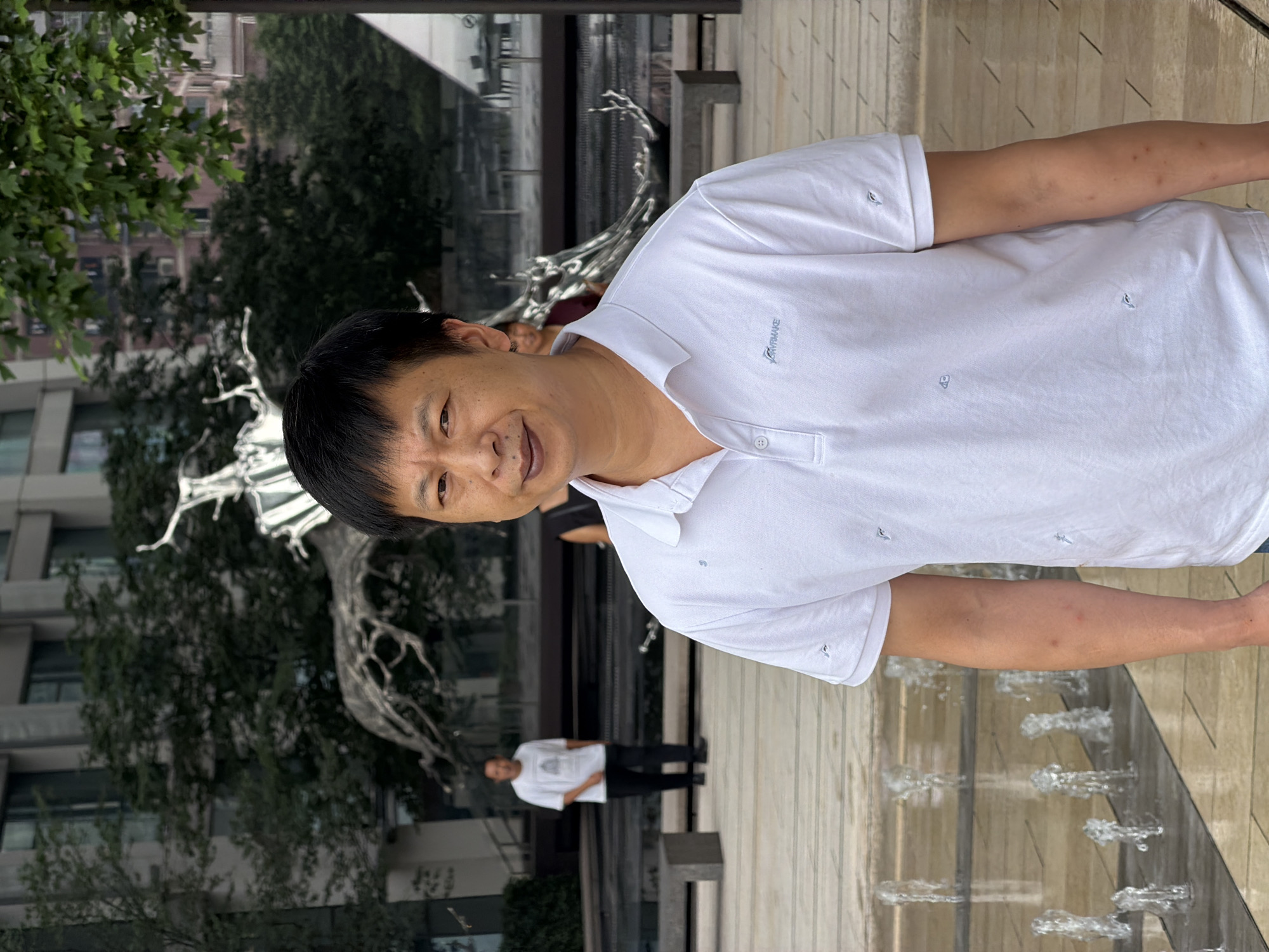A middle-aged East Asian man stands facing the camera in a bright, modern outdoor plaza with a water feature in the foreground. He is wearing a white polo shirt with a small embroidered logo on the left chest, and an embroidered dolphin motif repeated across the shirt. His hair is black and he has a neutral expression. In the background, a man in a white t-shirt and black pants walks away from the camera. Further behind him, a large, abstract metal sculpture is visible, set against a backdrop of modern buildings and trees. The plaza appears to be paved with light-colored tiles, and low concrete benches are scattered around. The overall environment suggests an urban public space, possibly a park or a courtyard in Yuzhong District, China.