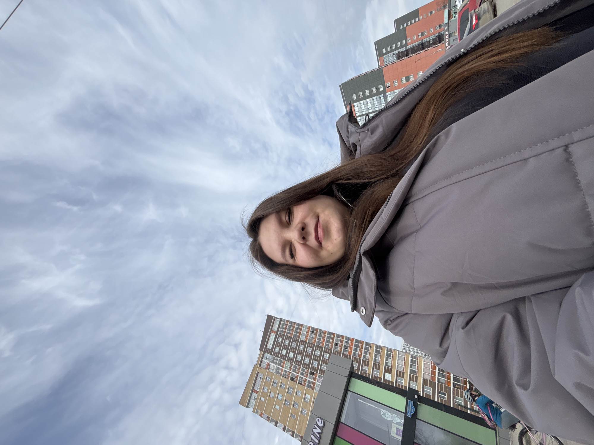 A selfie-style, tilted-angle photograph shows a young woman wearing a gray puffer jacket. She has long brown hair and appears to be outdoors. Behind her, partly obscured by the jacket and buildings, is a cloudy sky. In the background, multiple high-rise buildings with windows are visible. A portion of signage that reads "LINE" is seen on one of the buildings. Based on the building styles and signage, the location is identified as likely being in the city of Perm, Russia. A cyclist is visible at the bottom right of the frame.