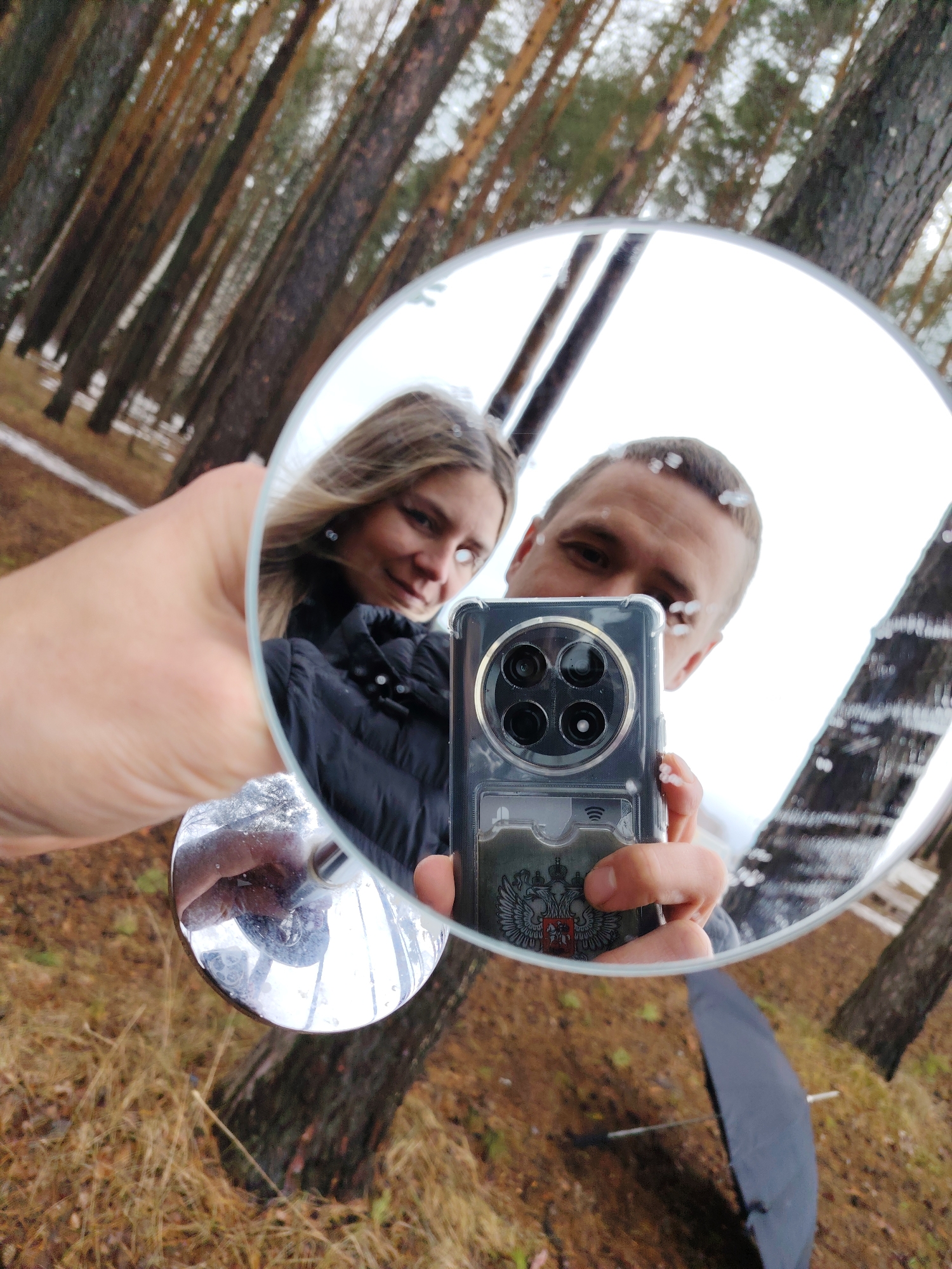 A mirrored self-portrait of a woman and a man holding a smartphone with a clear case that has an image of the Russian coat of arms is taken outdoors. They are in a wooded area near the city of Chaikovsky, Perm Krai, Russia. The trees in the background are tall and thin, with some snow on the ground. The woman is wearing a black jacket and the man appears to be wearing a grey shirt. The man is holding up a round mirror in order to take the photo. An umbrella is visible.