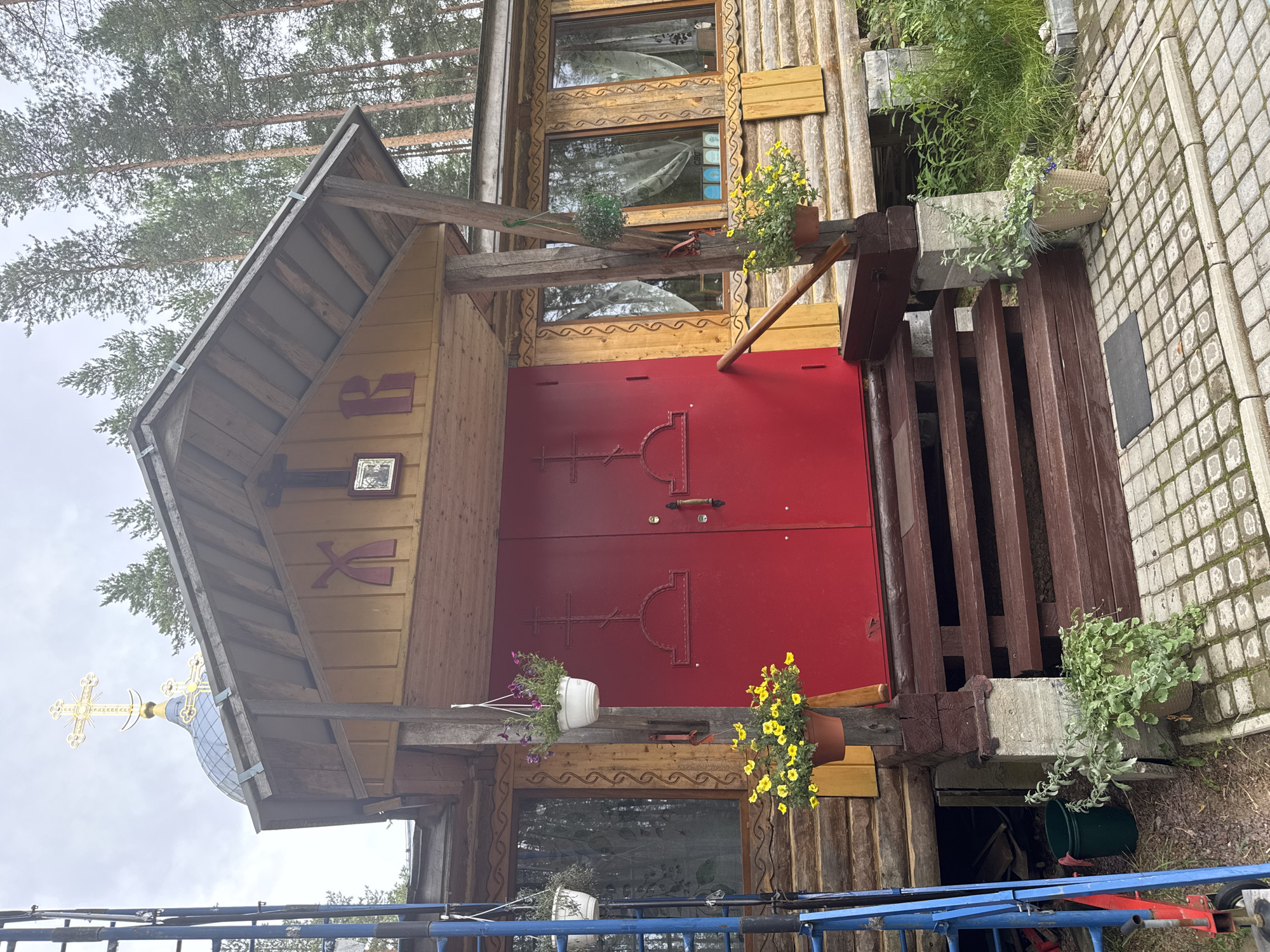 The image shows the exterior of a wooden church building in Nenyalki, Russia. The church has a red double door entrance with crosses on each door. Above the entrance, there is a wooden roof and a smaller decorative roof section with Cyrillic letters and a religious icon. The building is constructed of logs and has wooden window frames. Hanging flower baskets are placed at the entrance. The church is surrounded by trees, suggesting a rural or semi-rural setting. Scaffolding is visible on the left side of the image. The ground in front of the church is paved with cobblestones, and there are potted plants and grass in the foreground. The sky is overcast.