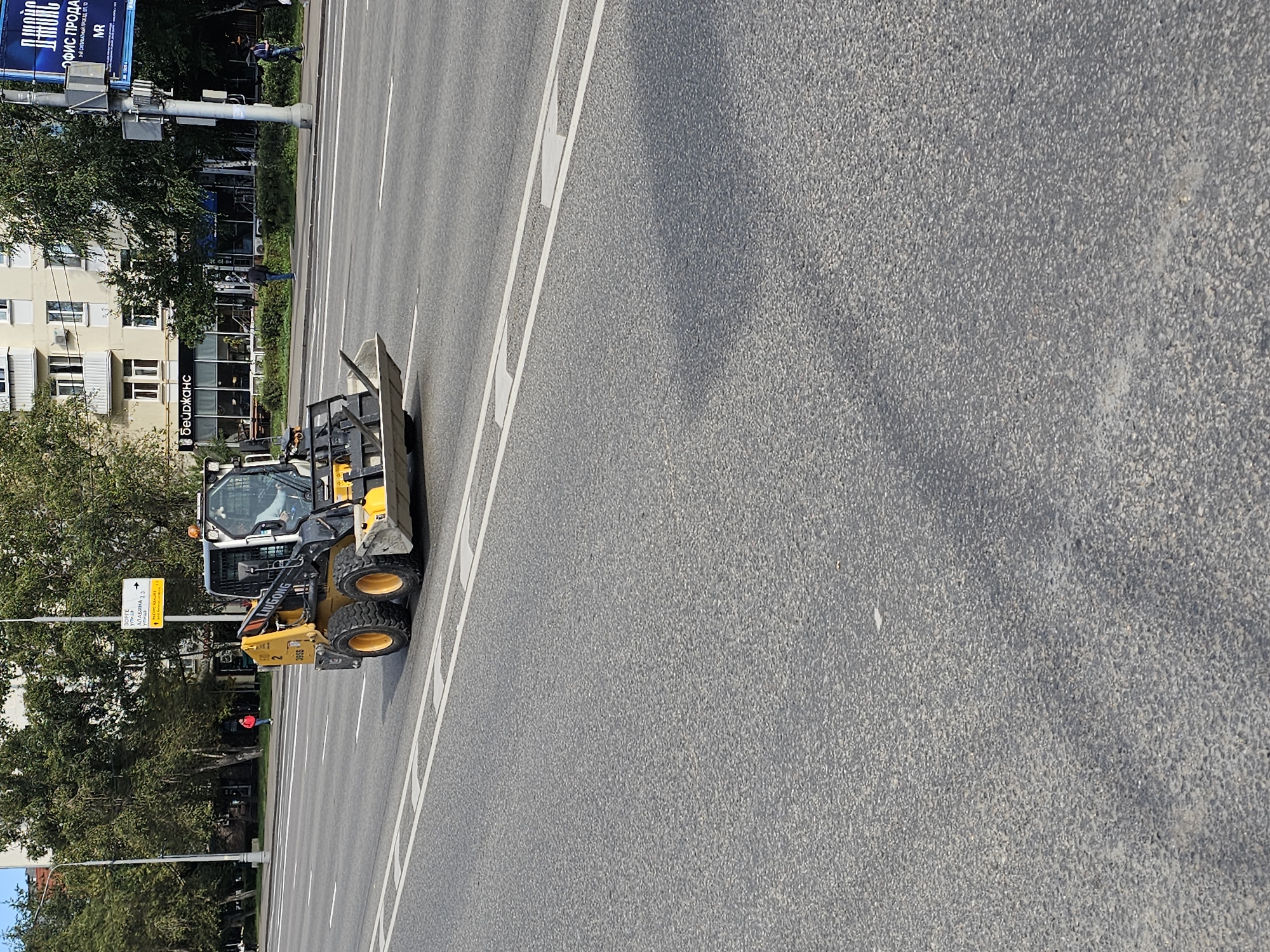 A yellow and black skid steer loader is driving on a paved road in Moscow, Russia. The skid steer loader has a bucket attached to the front and appears to be in operation. The road is a multi-lane street with traffic markings. In the background, there are trees, buildings, and street signs. The overall scene suggests an urban environment in Russia.