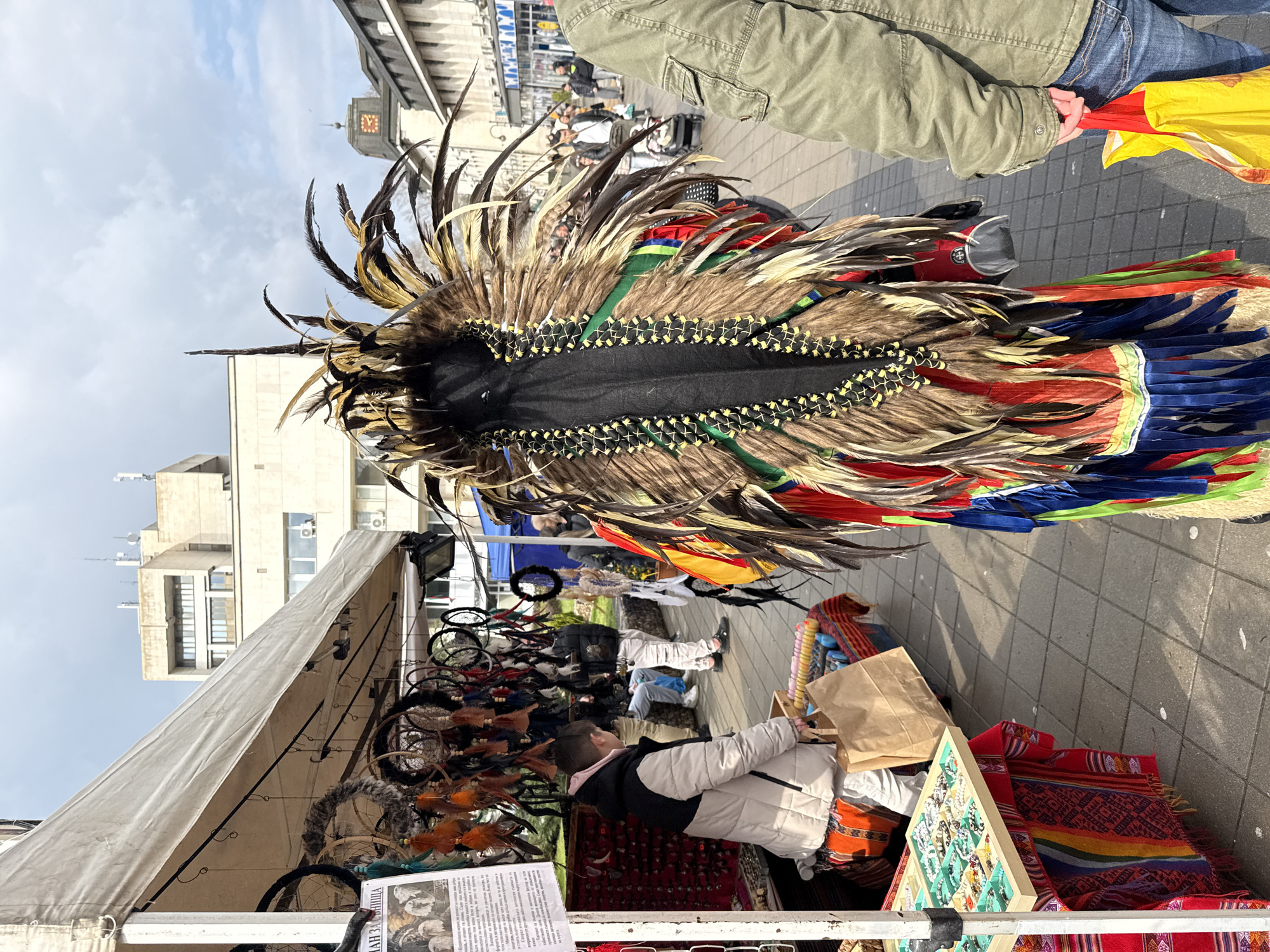 The image captures a bustling outdoor market scene in Lovech, Bulgaria. A person adorned in a large, elaborate feather headdress and multicolored clothing stands prominently in the foreground. The headdress features a black fabric base with rows of brown and gold feathers, accented by green and yellow decorative stitching.
Adjacent to the costumed individual is a market stall covered by a white canopy. Underneath the canopy, a vendor is present, sitting behind a display of various items, including dream catchers and jewelry. The vendor appears to be interacting with a customer, whose back is visible to the camera.
Beyond the immediate foreground, the urban environment of Lovech is visible. Buildings of varying heights and architectural styles line the street, and in the background, the city's clock tower can be identified. People stroll through the market, adding to the vibrant atmosphere. The street is paved with gray stone.