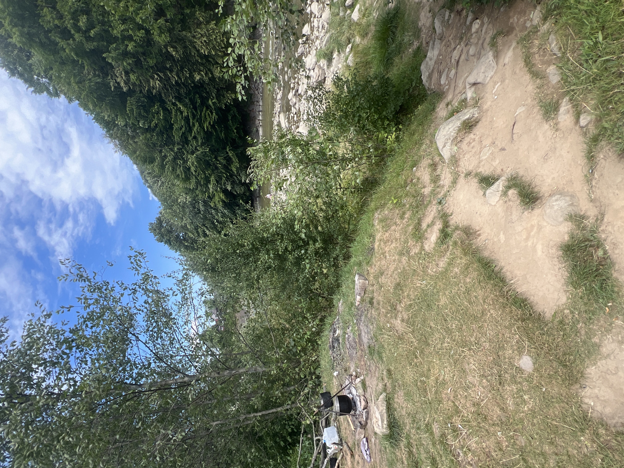 The image depicts a campsite near a riverbank in Yaremche, Ukraine. In the foreground, a campfire pit is visible with stones arranged around it, and a black cooking pot is suspended over the fire pit by a tripod. Scattered around the campsite are some discarded items, including a plastic container and a piece of cloth. The ground is a mixture of grass and dirt paths, with some large rocks. Behind the campsite, dense green trees and foliage line the bank of a shallow, rocky river. The river water is clear, with rocks visible beneath the surface. Further up the bank, more trees create a lush, natural backdrop. The sky above is blue with scattered white clouds. The overall scene suggests a rustic, outdoor setting, possibly a popular spot for recreation or camping in the Carpathian Mountains region of Ukraine.