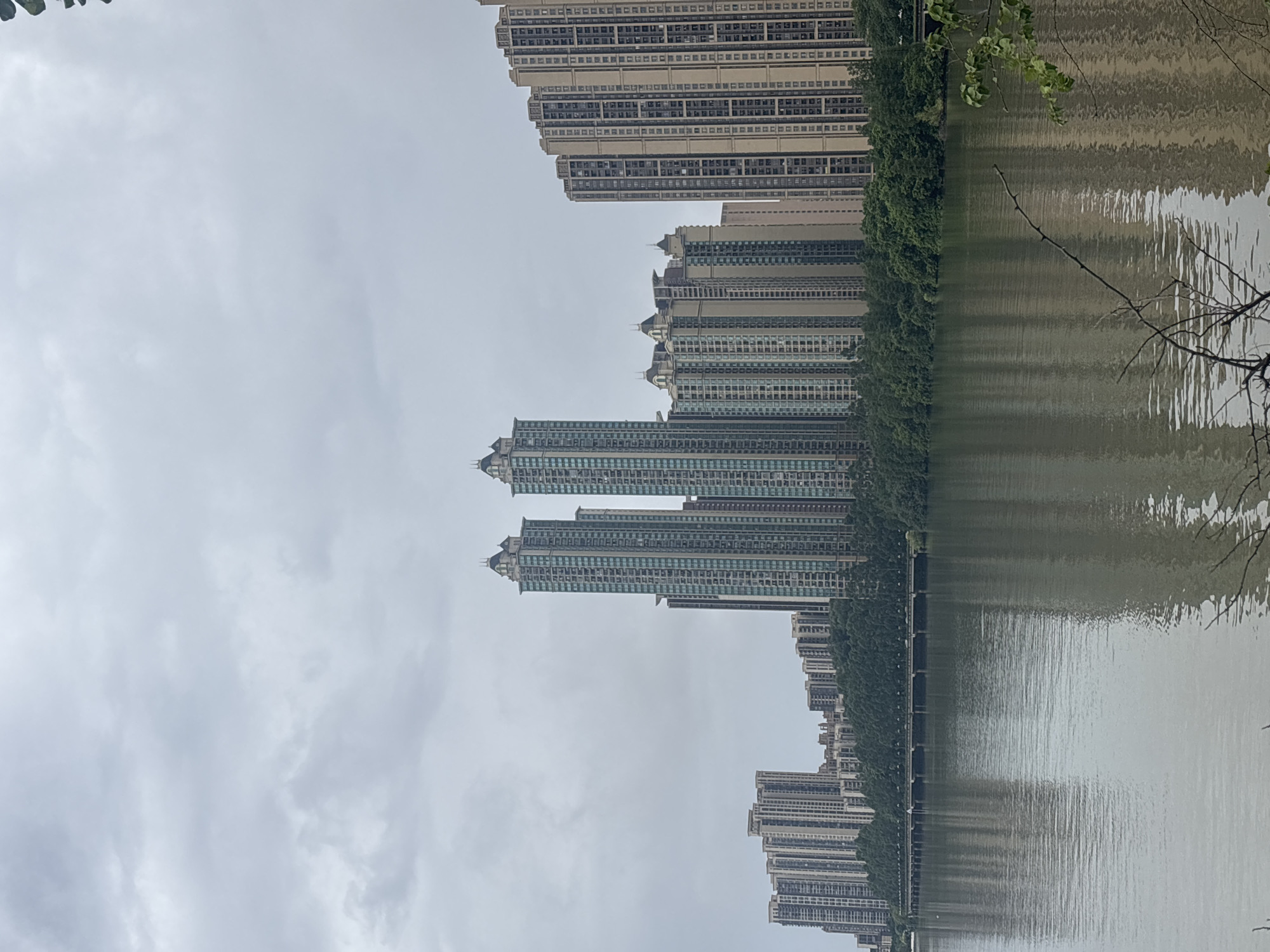 The image shows a panoramic view of a city skyline on a cloudy day. In the foreground, a large body of water reflects the sky and the buildings. The water appears murky and calm, with subtle ripples. Along the far bank of the water, a dense line of lush green trees creates a natural barrier between the water and the cityscape.
Rising above the trees is a series of modern high-rise apartment buildings. The buildings vary in height and architectural style, with many featuring repeating patterns of windows and balconies. Two particularly tall, slender towers with distinctive pointed tops stand out in the center of the composition. To the right, a longer, more uniform block of buildings stretches across the horizon. The architecture suggests a contemporary urban development. A long bridge, supported by numerous arches, is visible crossing the water in the distance, partially obscured by the trees. The sky is overcast with a uniform layer of gray clouds. The overall impression is one of a developed urban area situated alongside a natural waterway, under a subdued, cloudy sky. The scene is located in Nanhai District, China.
