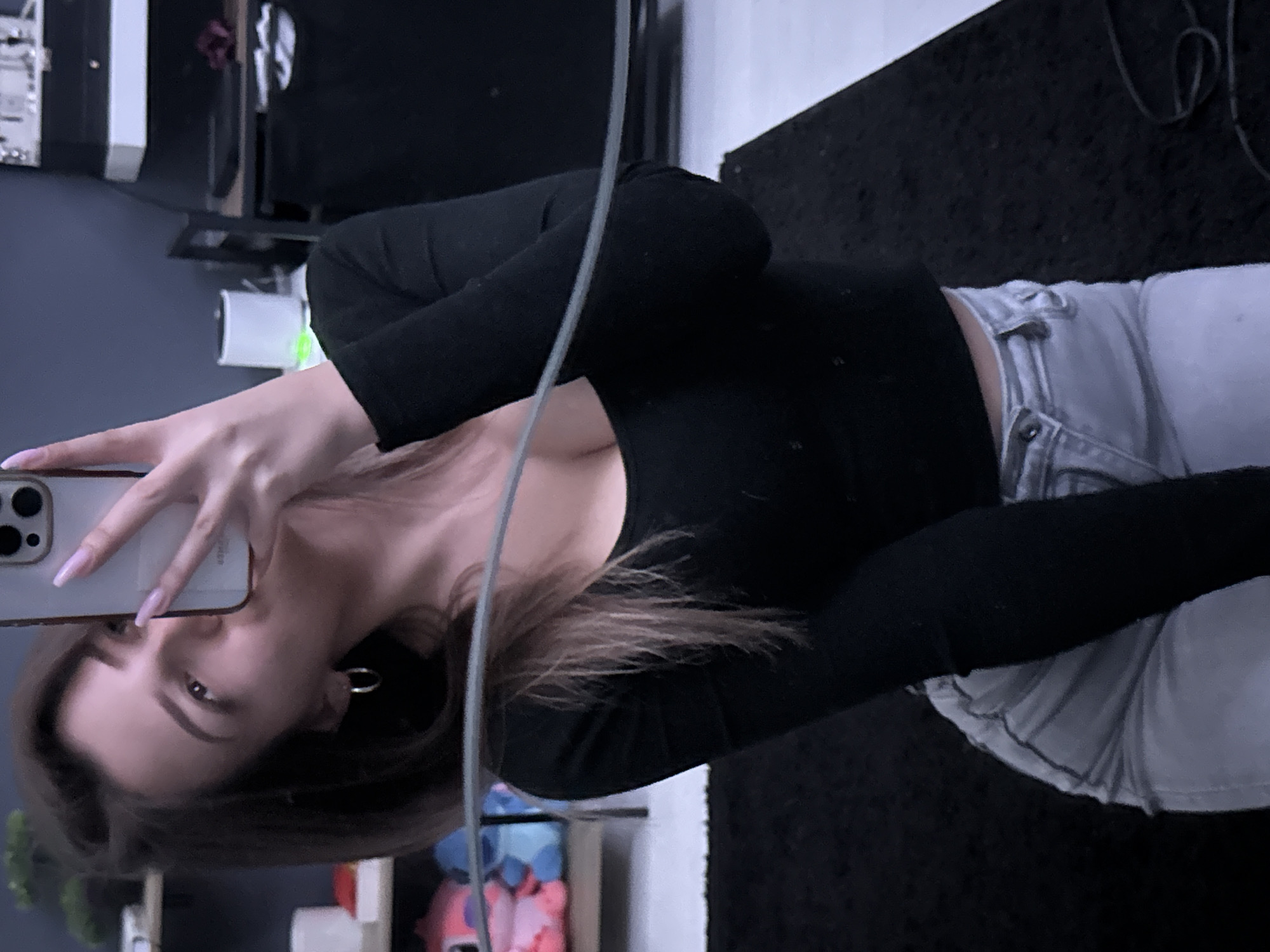 A woman is taking a selfie in what appears to be an indoor setting. She is holding a smartphone with a light-colored case, and her face is partially visible. She has brown hair, and her nails are painted a light color. She is wearing a black long-sleeved top and light-colored jeans. The background shows a room with a dark-colored rug and furniture, including a desk or shelf unit. There is also a reflection of objects like a monitor or computer tower in a glass surface. The presence of a wall and some scattered items suggests a private or residential space. There is no discernible location in the city of Sofia, Bulgaria.