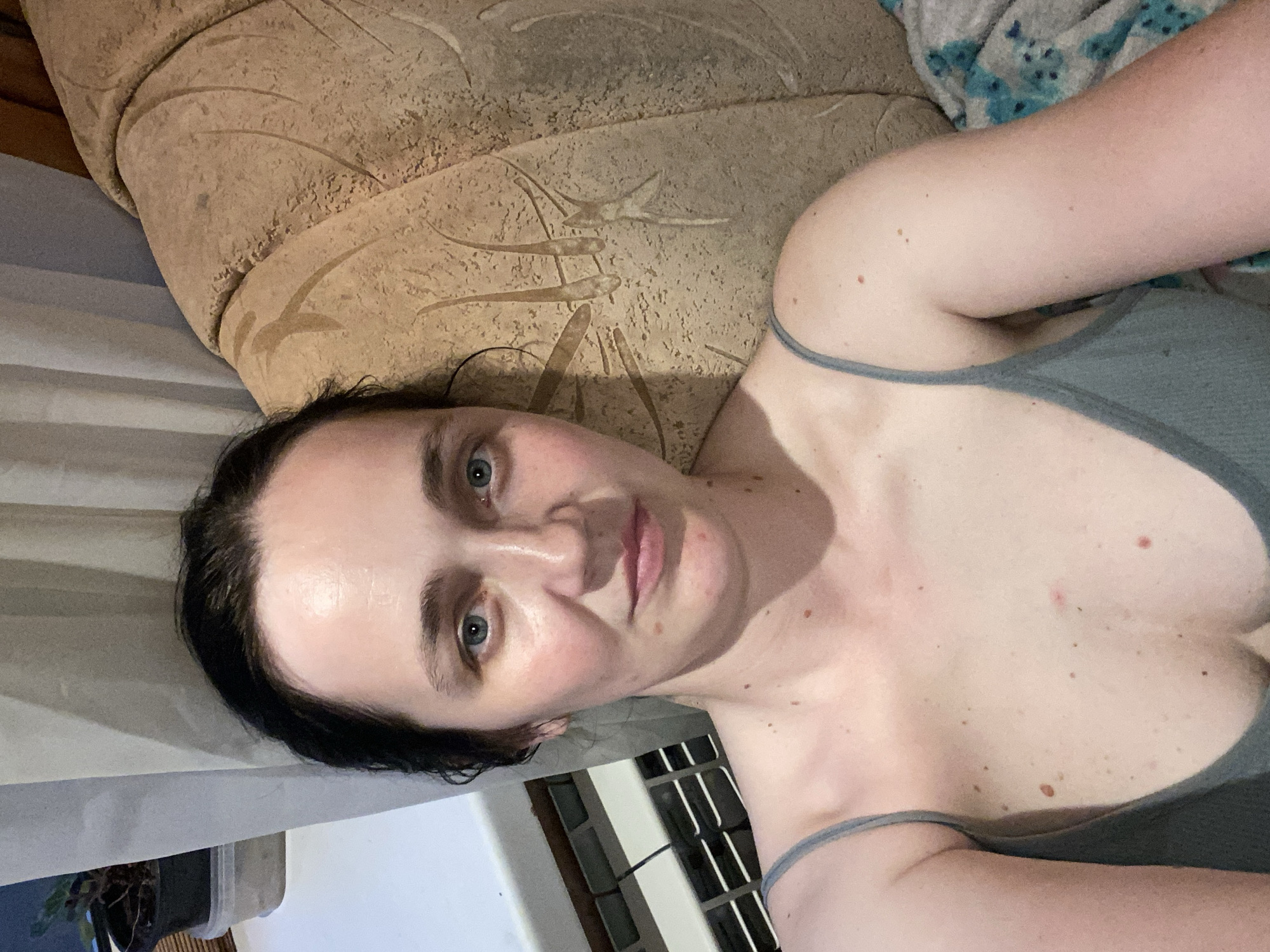 A Caucasian woman with dark hair, blue eyes, and fair skin is pictured from the chest up, looking directly at the camera. She is wearing a grey tank top. Her skin has several moles. She is seated on a textured, light brown couch with a blanket draped over it. To her left, a white curtain hangs beside a windowsill with a potted plant and a radiator. The background appears to be indoors.