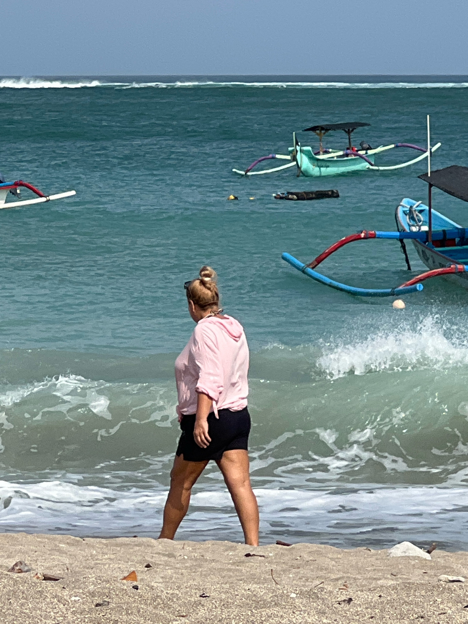 A full shot captures a woman standing at the edge of the water on a sandy beach in Kuta, Indonesia. She is facing away from the camera, towards the ocean, her blonde hair pulled up in a bun. She wears a pink hooded top and black shorts. Waves are crashing at her feet, and the water is a vibrant turquoise. Several traditional Balinese fishing boats, known as jukungs, are anchored in the sea. One boat, painted turquoise with purple and white outriggers, is in the mid-ground. Another blue and red boat is closer to the foreground. The sky is a pale blue with a hint of clouds. The beach is made of light-colored sand, with scattered debris visible.