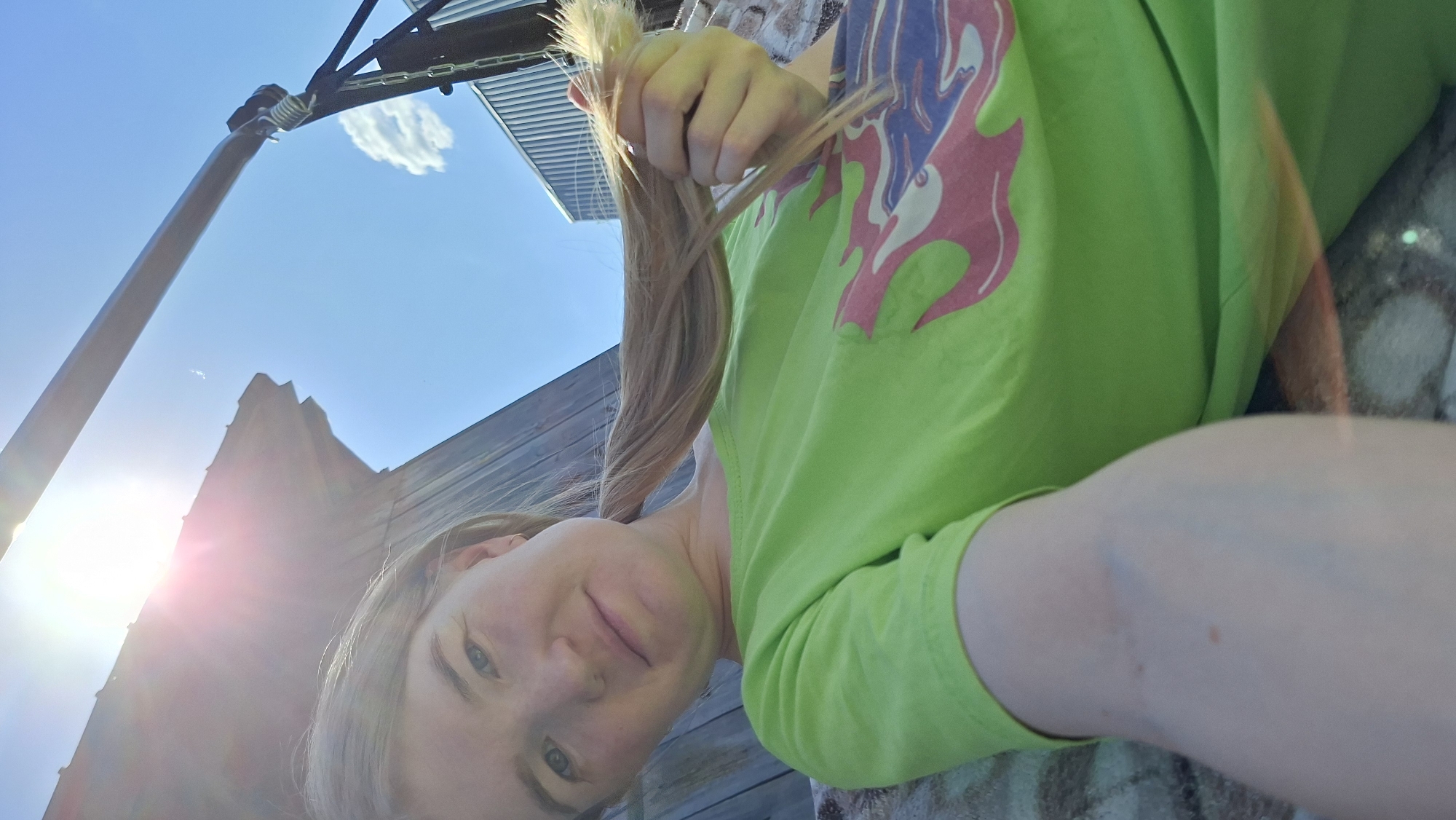 A selfie, taken at a low angle, shows a young woman with light skin and blond hair. She is looking at the camera with a slight smile. She is wearing a bright green t-shirt with a graphic design on the upper left chest. In her hand, she is holding a bunch of her own light blonde hair.
The background includes a bright blue sky with a few white clouds. Above her, there is a metal structure, possibly a basketball hoop. In the bottom left corner, there is a portion of a building that is likely located in the city of Большой Букор, Russia. The sunlight is strong and creating lens flare. The surface beneath the woman is textured, possibly a stone or patterned surface.