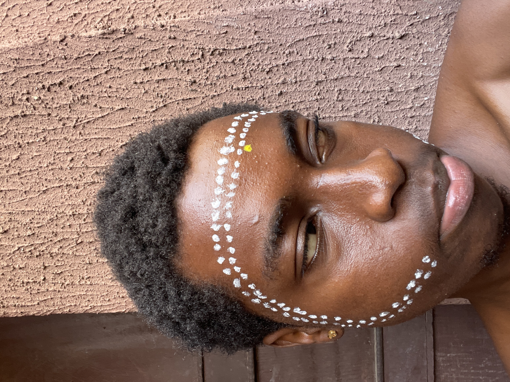 An individual with dark skin and short, dark curly hair is lying on a dark brown surface. A pattern of small white dots is painted on the individual's face, framing the forehead, cheek, and jawline on the right side. A single yellow dot is placed on the forehead. The individual is wearing a small gold earring in their left ear. The background consists of a textured, light brown surface. The setting is Nkpor, Nigeria.