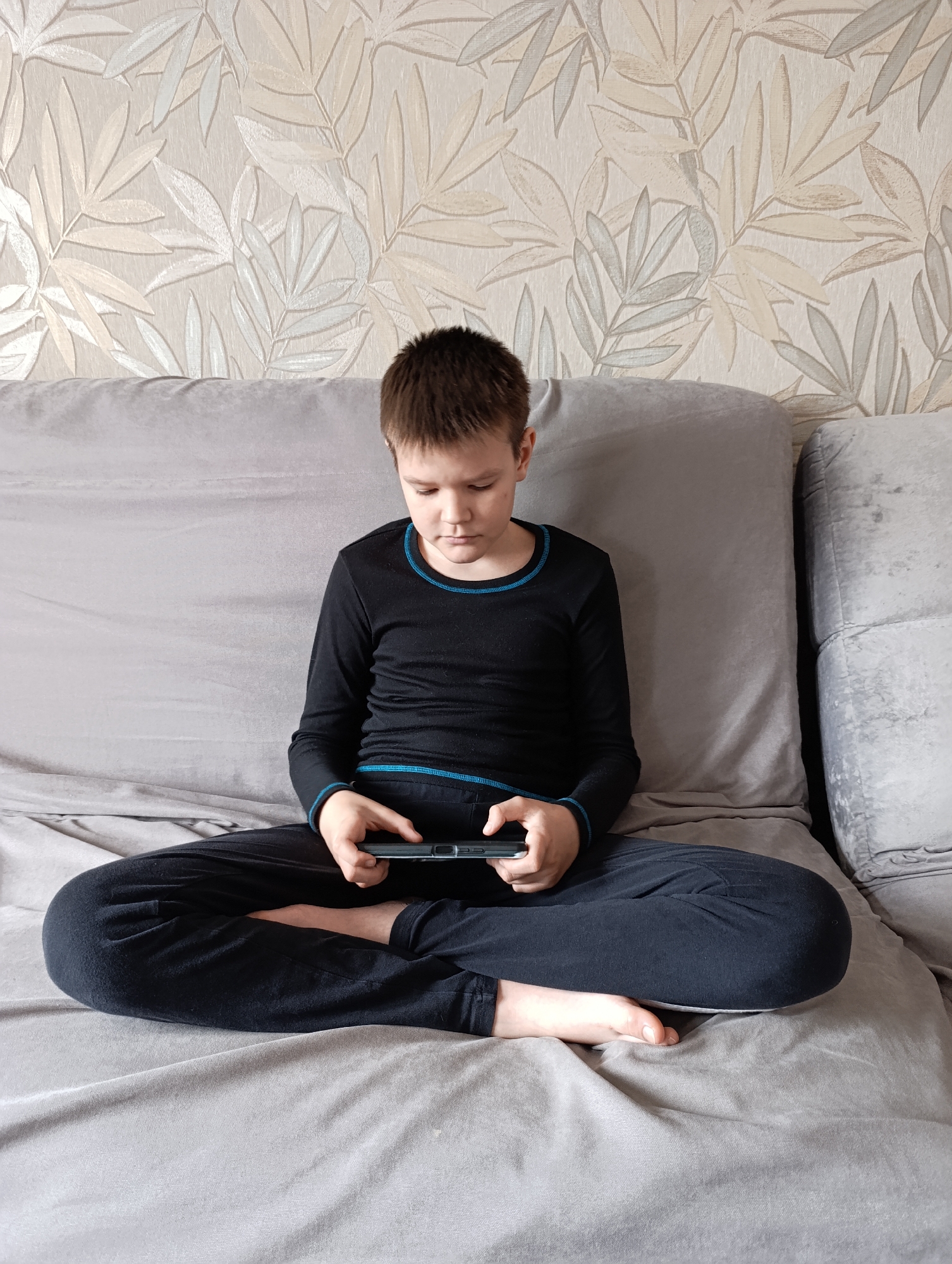 A young boy sits cross-legged on a grey sofa, looking down at a mobile device he is holding in his hands. He is dressed in a dark blue long-sleeved shirt and matching pants with a thin blue trim. The sofa is set against a wallpapered wall with a leaf pattern in beige and grey tones. The scene appears to be indoors. It's impossible to verify the location in Фоки, Russia, from the visual information provided.