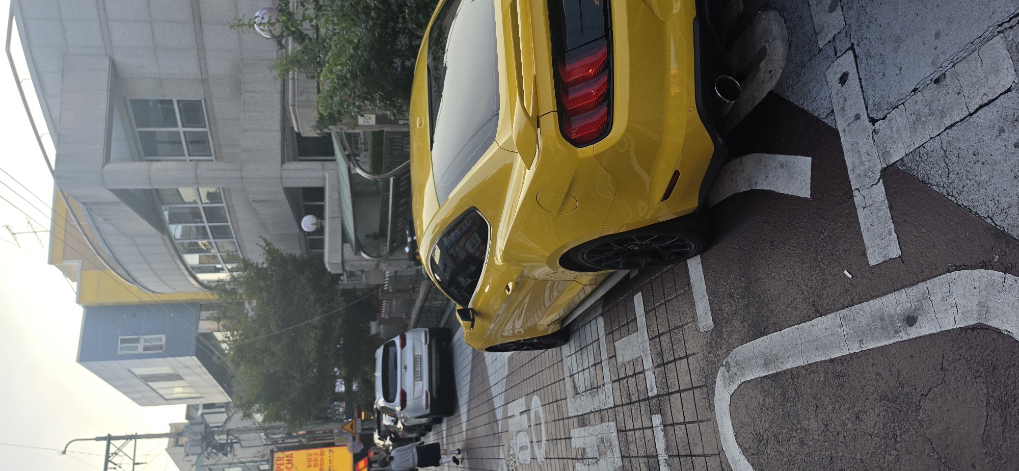 A yellow Ford Mustang coupe is parked on a street in Cheongju-si, South Korea, with its rear facing the camera. The car is parked on a brick-patterned sidewalk with white painted markings. In the background, a white SUV is parked behind the Mustang, and further down the street, a person with dark hair wearing a grey top and black skirt is walking away from the camera. Buildings line the street, one with a distinctive blue and yellow facade. Trees and streetlights are also visible. The sky is overcast.