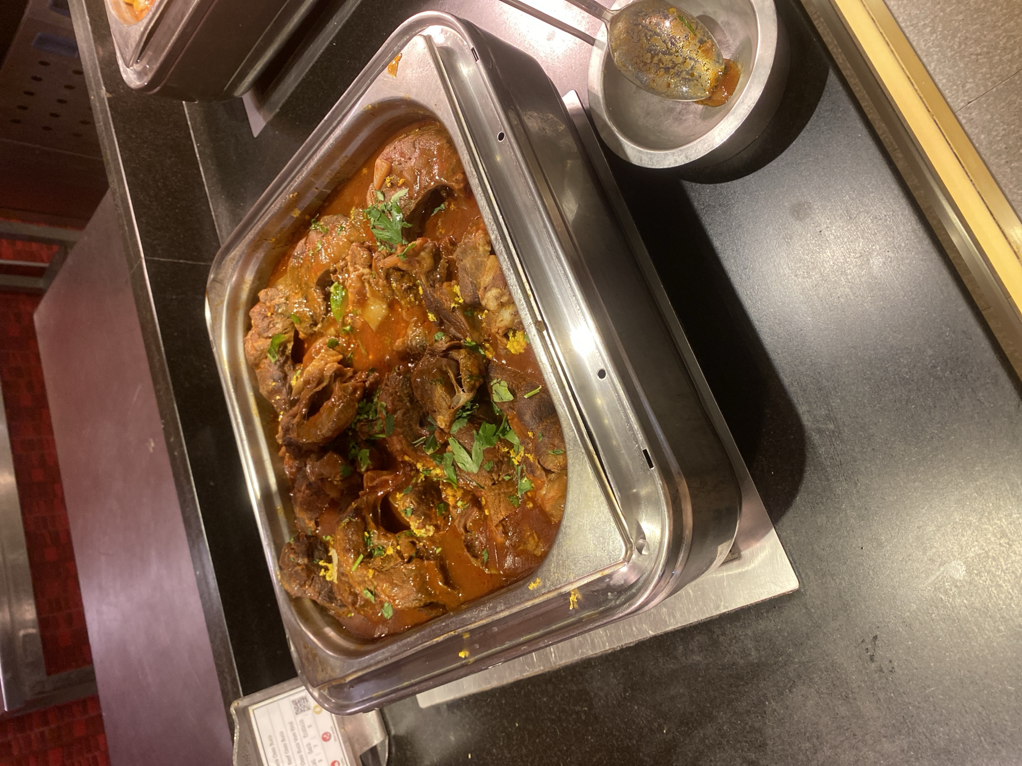 The image shows a close-up view of a buffet-style serving dish filled with braised beef in a rich, reddish-brown sauce. The beef appears to be tender and falling apart, garnished with chopped fresh green herbs, likely parsley, and flecks of yellow zest, possibly lemon or orange. The dish is presented in a rectangular stainless steel chafing dish, which is sitting on a metal stand. Beside the chafing dish, a small, shallow metal bowl contains a spoon with some of the braised beef and sauce on it, suggesting it has been used for sampling. The surface of the buffet counter is dark and speckled, and the background shows a red tiled wall and stainless steel elements, typical of a food service establishment. A small information card is partially visible on the left side of the image, listing menu items, including "Beef Osso Buco." The overall impression is of a warm, savory dish ready for serving. The location is identified as Aksu, Turkey, though the image itself does not contain any explicit visual cues to confirm this specific location.