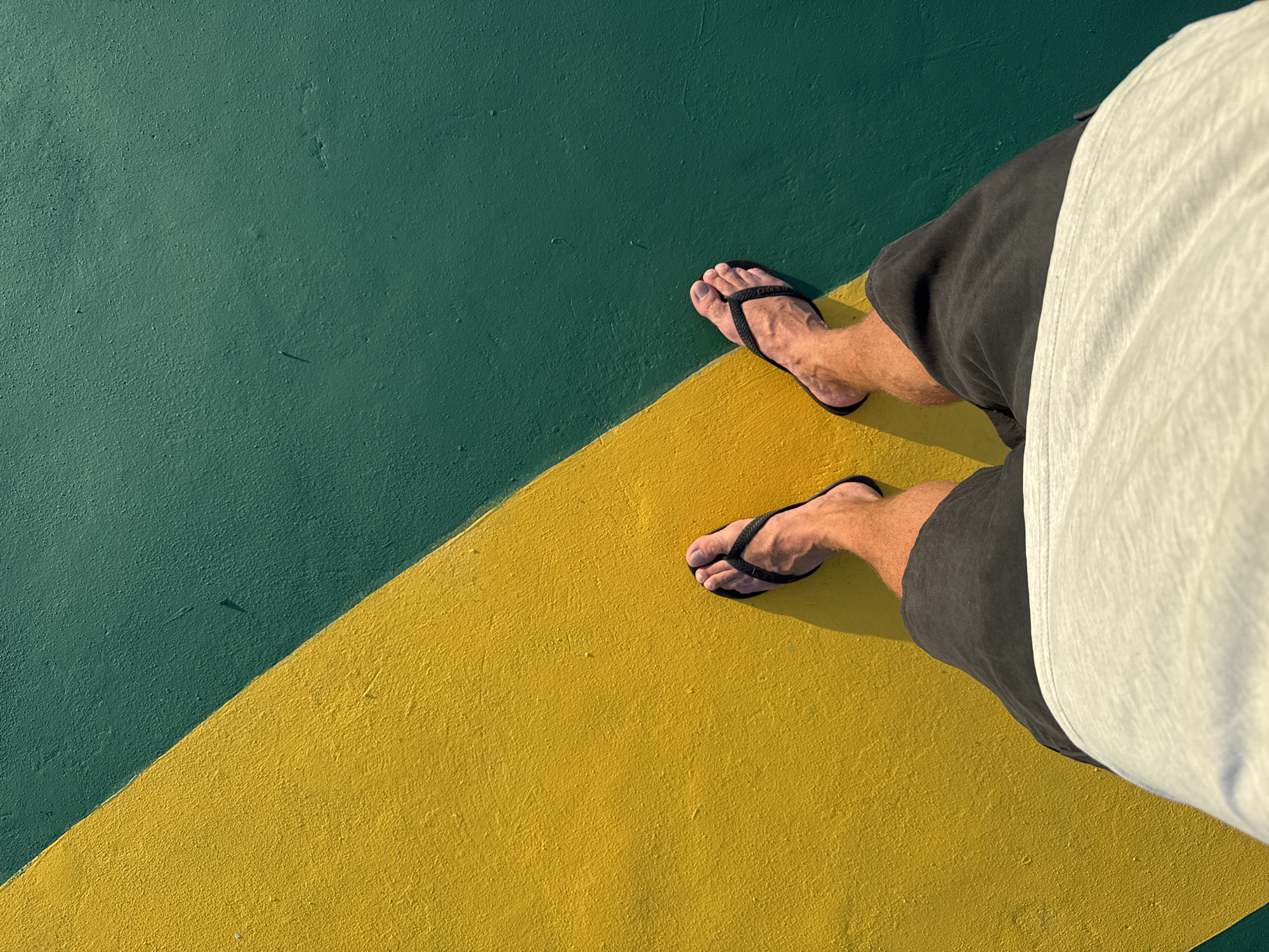 A high-angle shot depicts a person standing on a surface divided diagonally into green and yellow sections. The person is wearing black flip-flops, dark gray shorts, and a white shirt that is partially visible. The surface texture appears to be rough. The location is inferred to be Pasir Panjang, Indonesia.