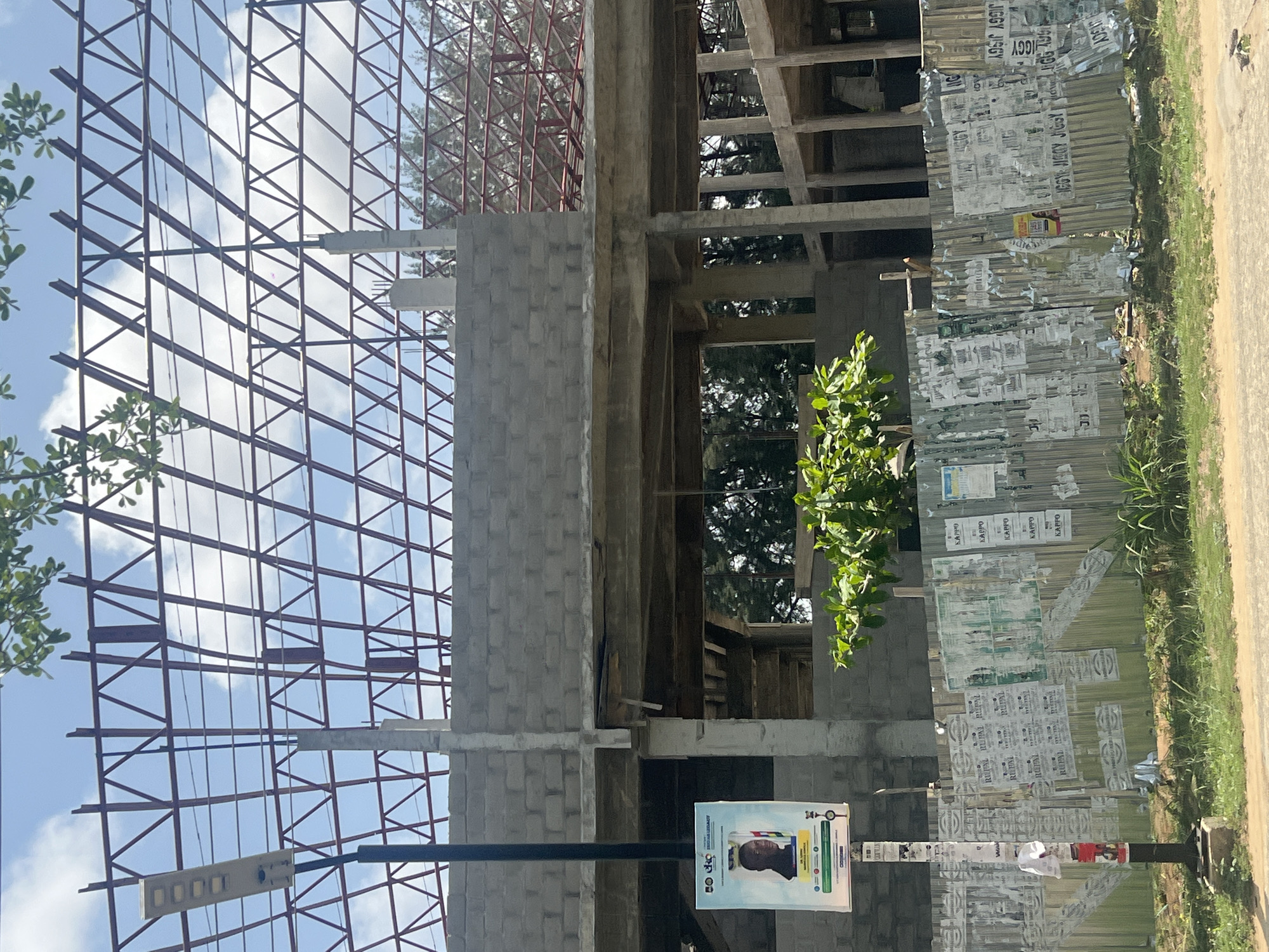 The image depicts a construction site in Ijanikin, Nigeria. An unfinished multi-story building stands prominently, its skeleton of concrete blocks and steel framework exposed. Scaffolding and support structures are visible within the building's open spaces. A metal fence, covered with posters and advertisements ("RUPAL", "KAPPO", "JIGGY JISGY"), runs alongside the construction site. A street light pole with a political advertisement attached is in the foreground. Vegetation is visible on the periphery of the scene, including a small tree in front of the building and grass along the side of the road. The sky is partially cloudy.