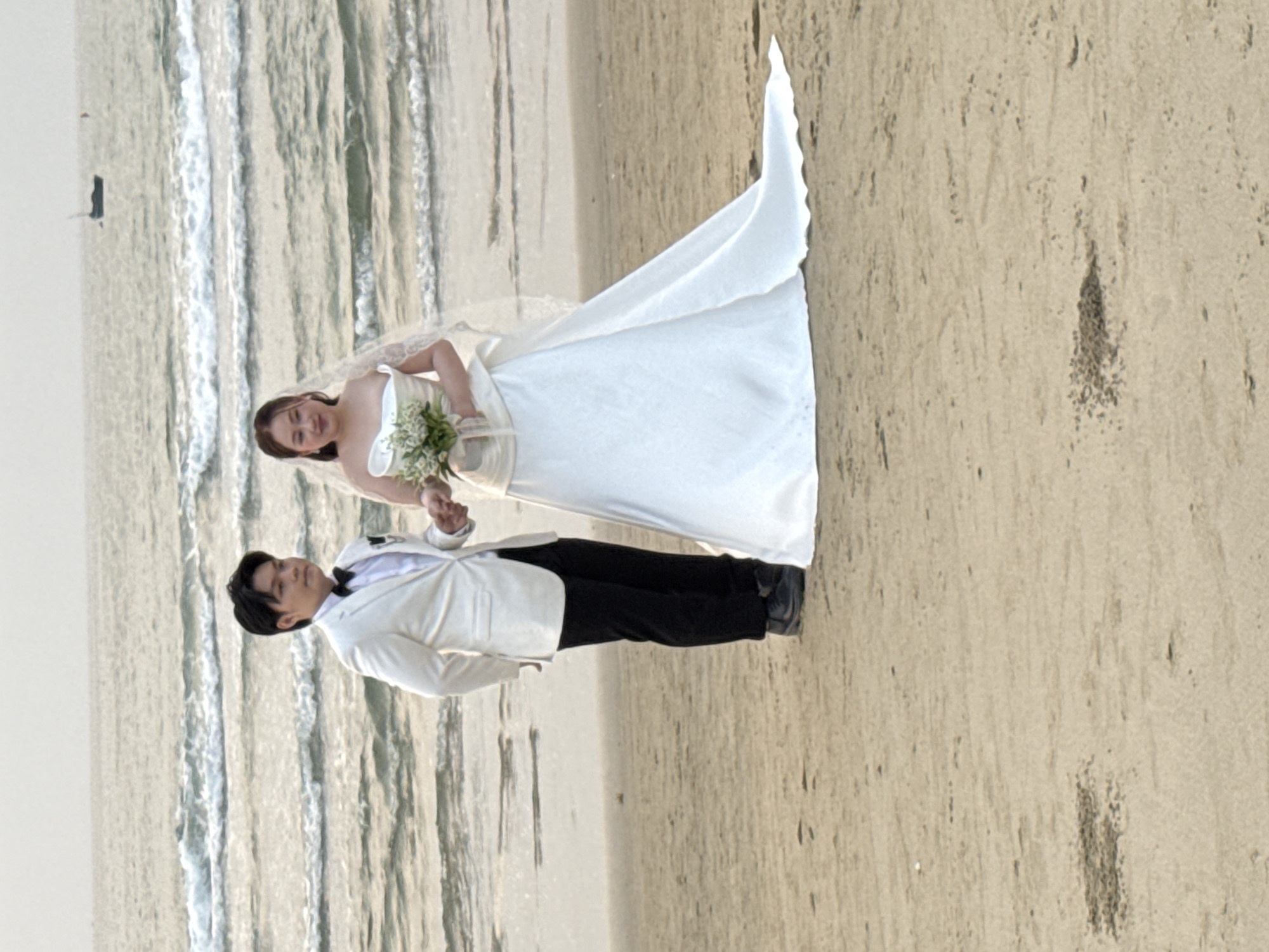 A wedding couple is posing on a beach in Đà Nẵng, Vietnam. The bride is wearing a white wedding gown and veil, and holding a bouquet. The groom is wearing a white jacket and black pants. They are holding hands. The background includes the ocean and the beach.
