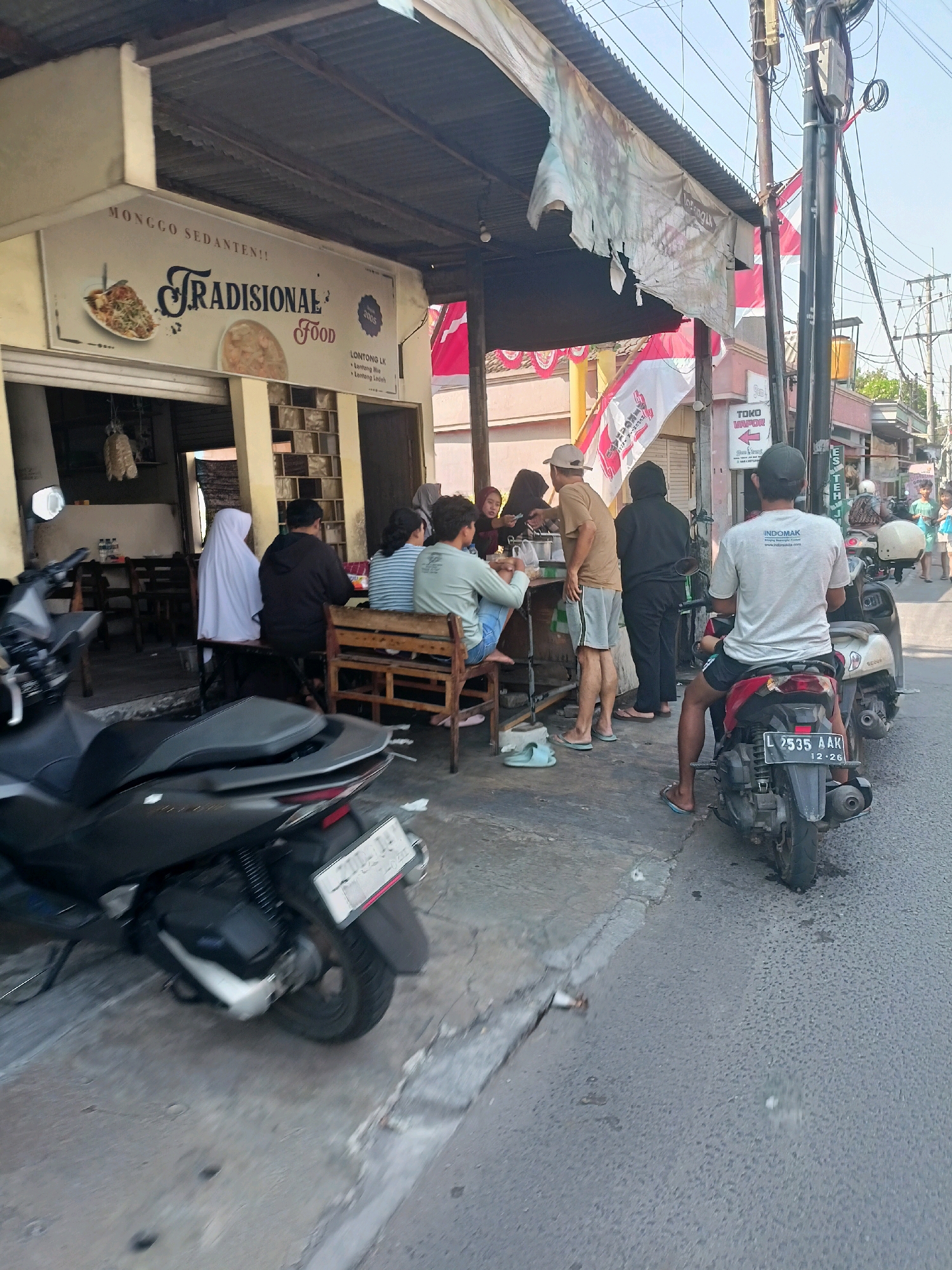 This is a medium shot, taken from ground level, of a street food stall and its surroundings in Surabaya, Indonesia. The stall, named "TRADISIONAL FOOD", is covered by a corrugated metal roof supported by wooden posts. A sign above the entrance displays images of food and the text "MONGGO SEDANTEN!! TRADISIONAL FOOD". Several people are seated at tables under the shelter, some eating and some conversing. One person in the foreground is on a motorcycle, facing away from the camera, with a license plate that reads "L 2535 AAK 12-26". Other motorcycles are parked nearby. In the background, a red and white Indonesian flag is visible, along with power lines and a "TOKO VAPOR" (vape shop) sign. The overall environment appears to be a busy, open-air market or street.