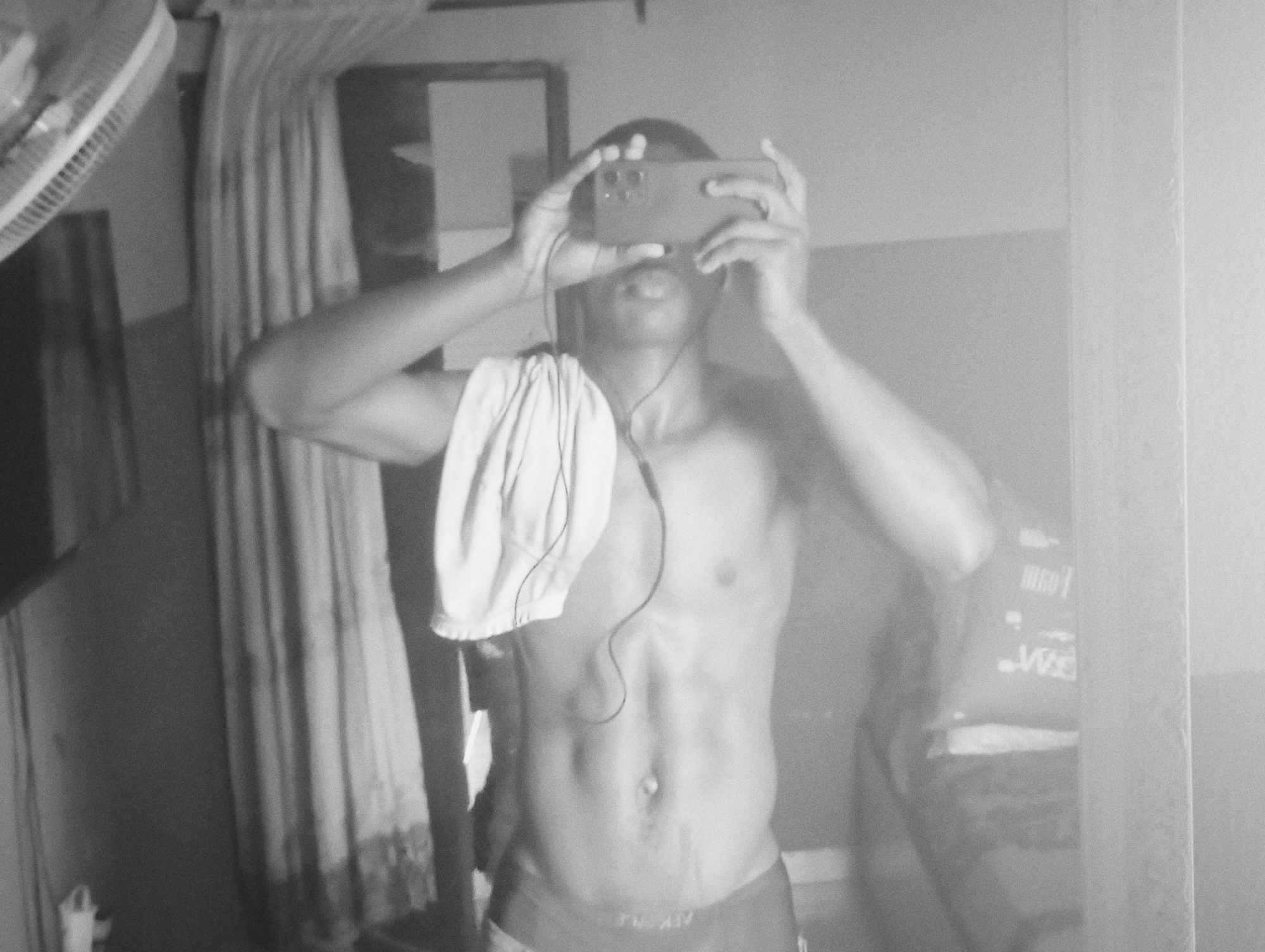 A black and white medium shot depicts a shirtless man with dark skin holding a smartphone up to obscure his face. He stands indoors in front of a mirror, capturing his reflection. The man's physique is toned, with visible abdominal muscles. A white towel is draped over his left shoulder. He wears dark-colored briefs. Earbuds hang down his chest. The backdrop includes a partially visible curtain on the left side of the frame, a dark window frame behind him, and what appears to be furniture or a bed reflected in the mirror on the right. Due to lack of data, a specific location within Nkpor, Nigeria cannot be identified.