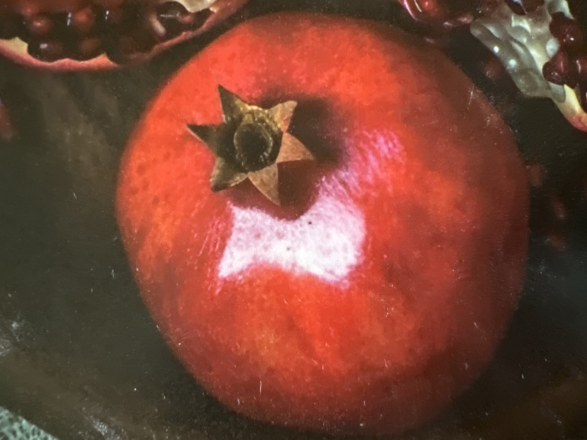 The image is a close-up, top-down view of a single, ripe pomegranate. The pomegranate is a vibrant red with a subtle gradient of lighter red and orange on its surface, indicating ripeness. A star-shaped calyx, characteristic of pomegranates, is visible at the top, exhibiting a brown and green hue. A bright, white reflection is prominent on the left side of the fruit, suggesting a light source in the environment. To the right of the main pomegranate, a portion of another pomegranate, split open, is visible, revealing its juicy, ruby-red arils. The background is dark and appears to be a textured surface, possibly a tabletop or dark fabric. The lighting is soft, creating shadows that add depth and dimension to the scene. There is no discernible location in the city of Minsk, Belarus, or any people or actions depicted in this image.