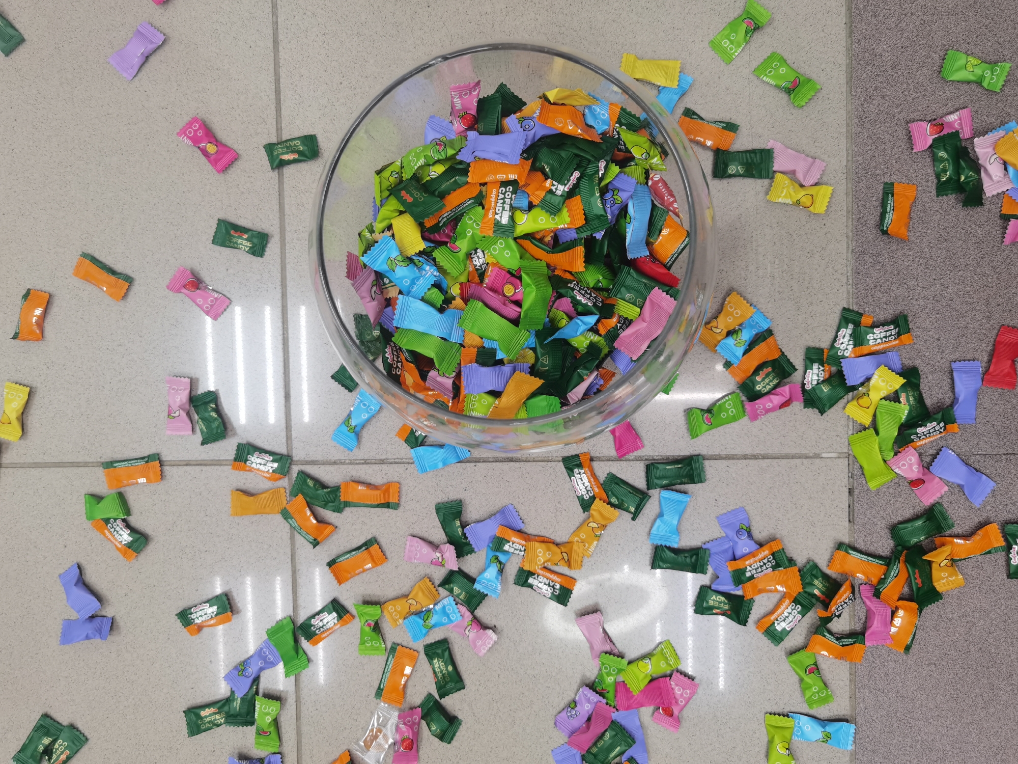 The image shows a large glass bowl filled with colorful wrapped candies sitting on a tiled surface. The tiled surface is light gray and has a grid pattern. Many of the same candies are scattered around the bowl on the tiled surface. The candies are wrapped in various colors including orange, green, pink, blue, yellow, and purple. The wrappers have writing and images on them, including the words "COFFEE CANDY". The candies are various flavors like Cappuccino, Mint, Strawberry, Peach, Lemon and Blueberry. The overall scene gives the impression that the candies have spilled or been scattered from the bowl. The location cannot be determined.