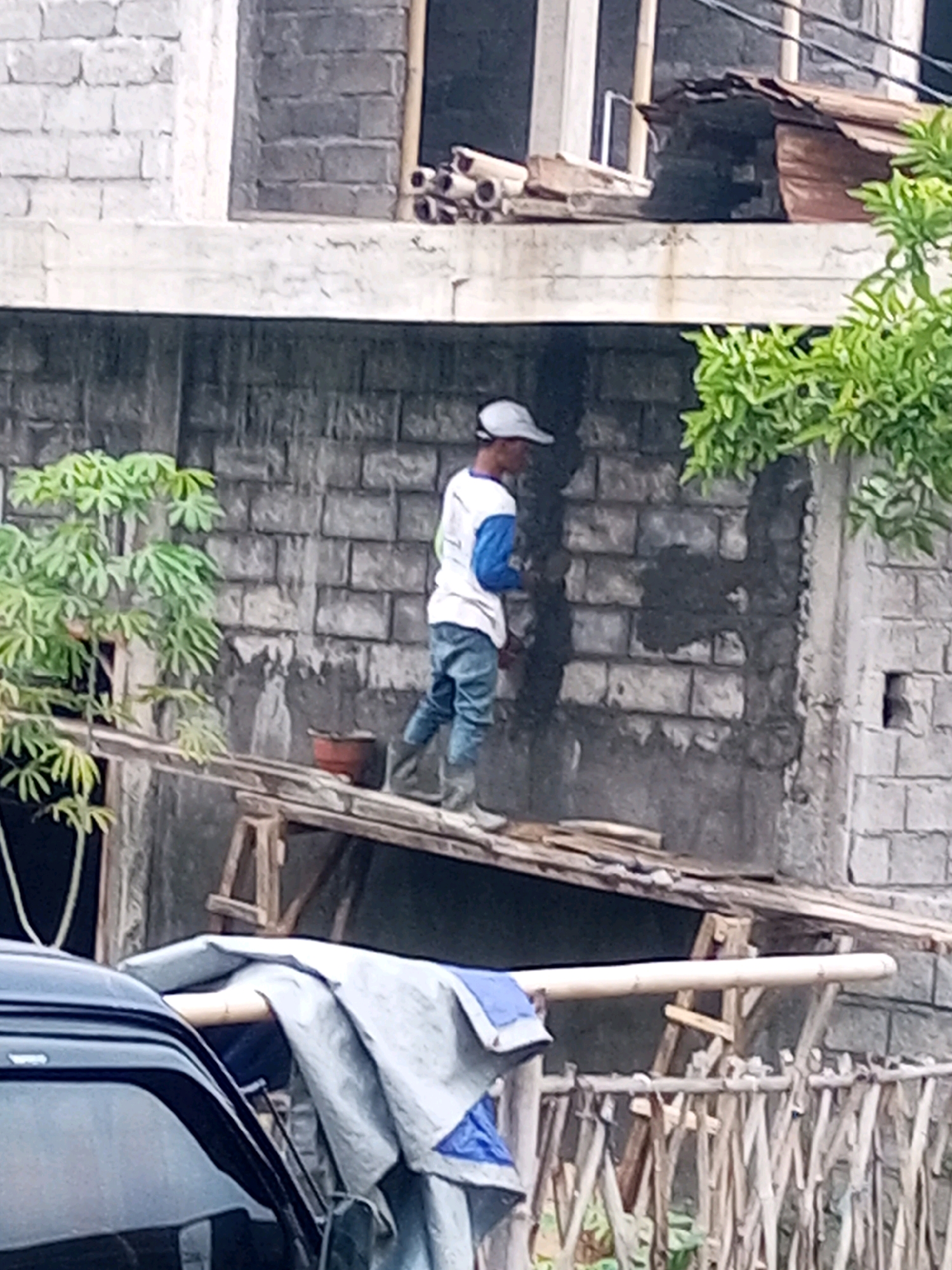 A construction worker stands on a wooden platform in Andongsili, Indonesia, applying plaster to a brick wall. The worker, wearing a white shirt with blue sleeves, blue jeans, and a white cap, is facing away from the camera. A terracotta pot and a stack of bamboo poles are also visible on the platform. In the background, a dark-colored vehicle is partially visible, covered with a tarp. The environment appears to be a residential area undergoing construction, with visible scaffolding and building materials. The overall scene depicts a typical day of manual labor at a construction site.