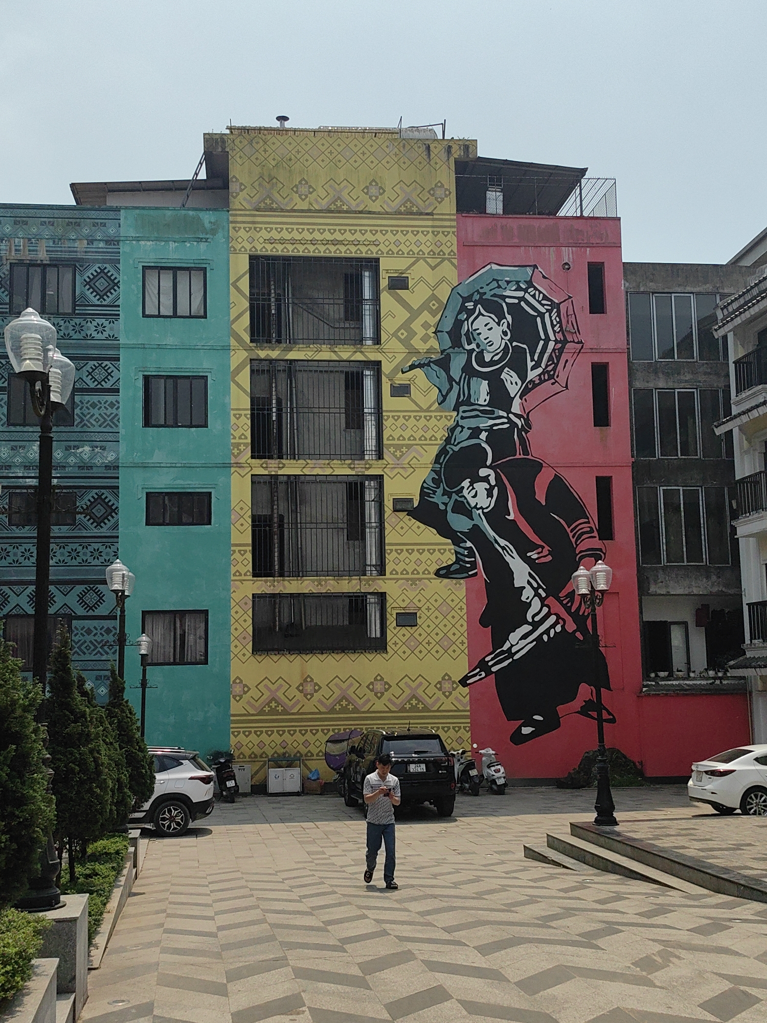 The image displays an urban street scene in Sa Pa, Vietnam, centered on a large building facade. The building is segmented into three vertical sections: a teal-colored section on the left, a yellow section in the middle, and a pink section on the right. Both the teal and yellow sections are adorned with repetitive geometric patterns. A large mural, primarily in black, white, and teal, covers parts of the yellow and pink sections. This mural depicts two stylized figures in traditional attire; the upper figure holds an open umbrella while the lower figure is positioned beneath.
In the foreground, a paved plaza area with a chevron-like pattern in light and dark gray extends from the bottom of the frame towards the building. A single male individual walks towards the viewer from the middle ground, looking down at a white smartphone held in both hands. He is wearing a light-colored, short-sleeved patterned shirt, blue jeans, and dark sandals.
To the left, a series of green conical shrubs are planted along a raised curb, adjacent to a dark lamppost with a clear, multi-faceted glass fixture. Another similar lamppost is visible further back. Vehicles parked near the building include a white SUV on the left, a black SUV, a white scooter behind the black SUV, and a partially visible white sedan on the far right. The sky is clear and bright, indicating daytime conditions.