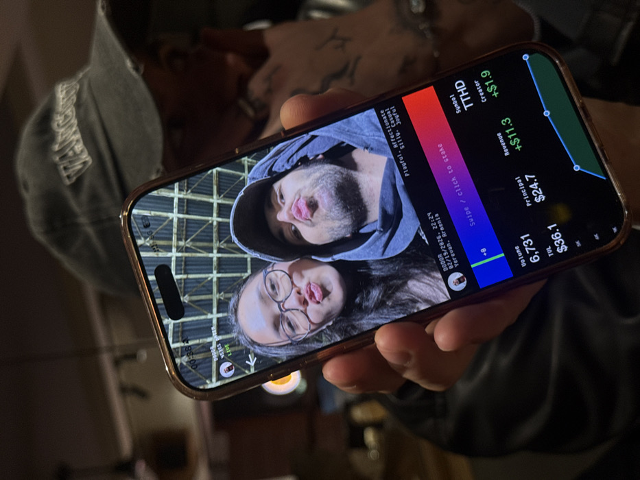 A hand holds a smartphone displaying a screenshot of a social media interface. A selfie of a man and a woman is in the background. The interface shows: live video from Yerevan, Armenia; "DODOR"; and the date as 02/19/2026, 22:24. The video's status is "Playful, Affectionate, Silly, Casual, Joyful". On the right side, the interface displays a financial chart, including "Volume 6,731", "TUL $36.1", "Principal $24.7", "Revenue +$11.3", "Symbol TTHD", and "Creator +$1.9".