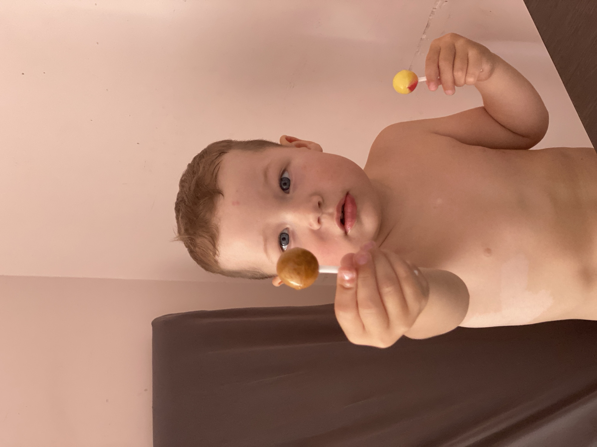 The image shows a young child with blue eyes and light brown hair lying down. They are holding two lollipops, one in each hand. The lollipop in the child's right hand is brown, while the one in their left hand is yellow with a red tip. The child appears to be looking at the camera with a neutral expression. The background is a plain pink wall. The child is shirtless, and their skin appears slightly damp, suggesting they may have recently been in water, perhaps a bath or pool. A dark surface is visible in the lower portion of the image, possibly a towel or mat. The lighting is soft, and it's difficult to determine the time of day or weather conditions. No visible text is present in the image. The location context of Sochi, Russia, does not provide specific visual cues within the image itself.