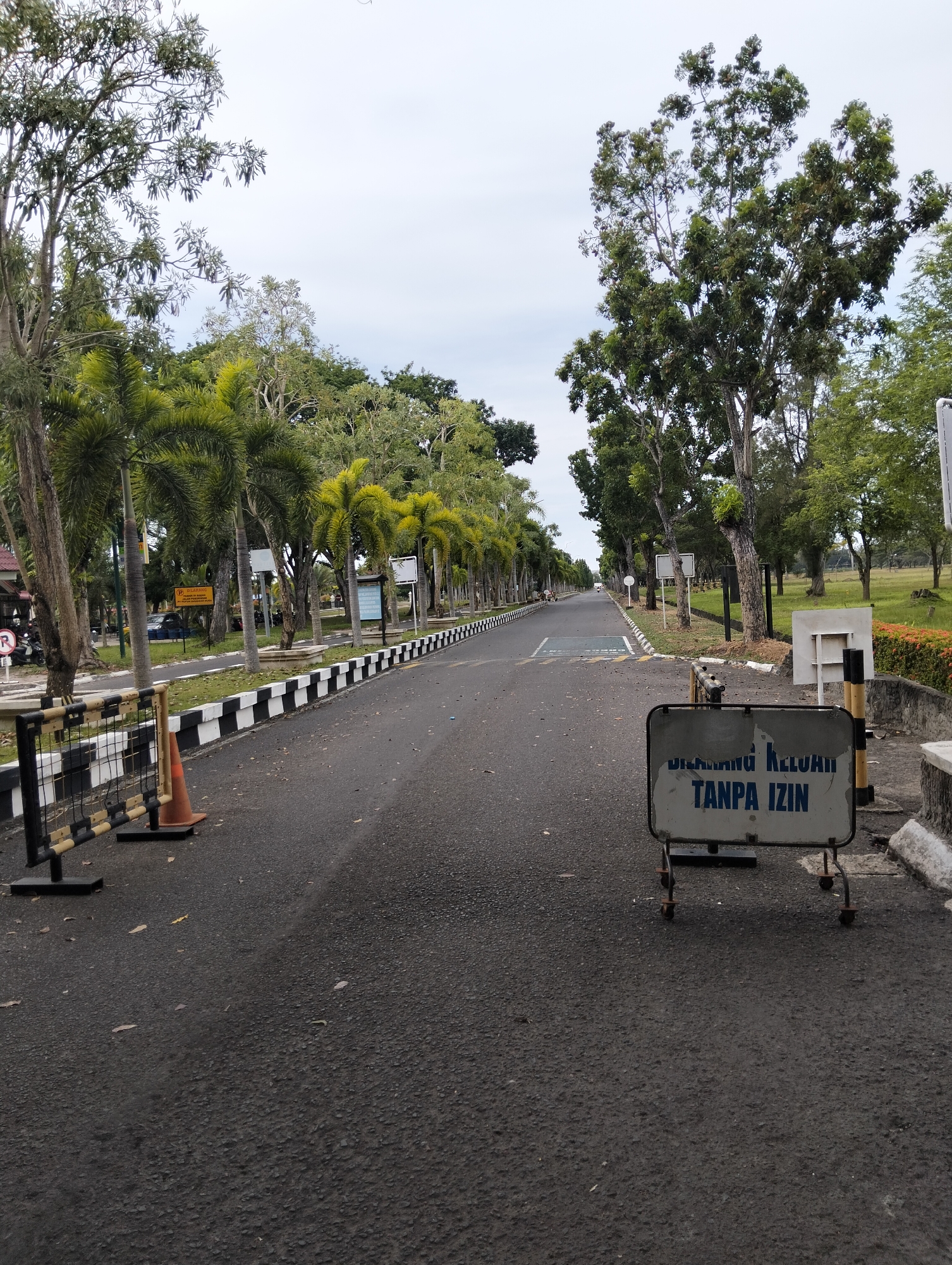 The image shows a straight, paved road lined with trees on both sides. In the foreground, a black and yellow barricade with a safety cone is positioned on the left side of the road. On the right side, a metal sign with blue lettering on a white background reads "DILARANG KELUAR TANPA IZIN," which translates to "Forbidden to Exit Without Permission." Several palm trees and other deciduous trees are visible along the road. The sky is overcast. The scene appears to be in a park-like or institutional setting, possibly a campus or a guarded facility. The location is identified as Tambontunong, Indonesia.
