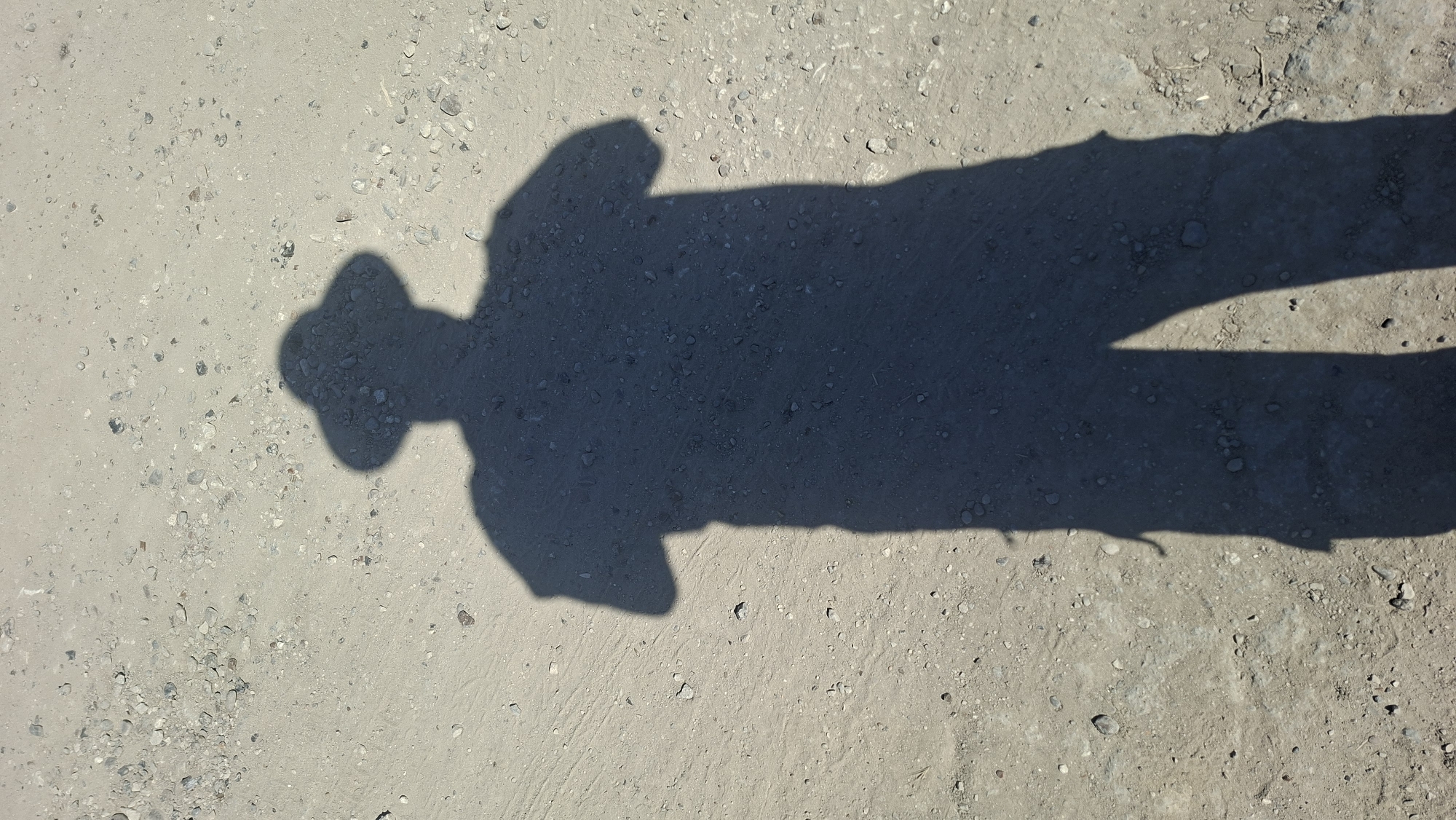 The image shows the shadow of a person wearing a hat and standing on a dirt ground with small pebbles. The shadow is cast on a sandy, uneven surface with scattered rocks and debris. The lighting suggests it is daytime, with the sun likely to the left and slightly behind the subject, creating a long shadow. The background is the ground itself, with no identifiable buildings or landmarks present. No specific details about Hîncești, Moldova are visible in the image.