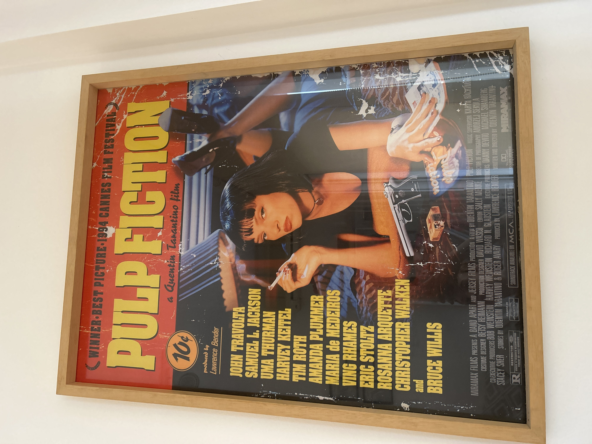 The image shows a framed movie poster for "Pulp Fiction" hanging on a white wall. The poster features the film's title prominently, along with a portrait of Uma Thurman as Mia Wallace. The poster is worn and appears to be aged, with some damage to the edges. The frame is made of light-colored wood. The wall is plain and white, with no other visible decorations or features. There is no discernible indication of the location being Buenos Aires, Argentina, or any other specific city. The scene is static and depicts a framed object indoors.