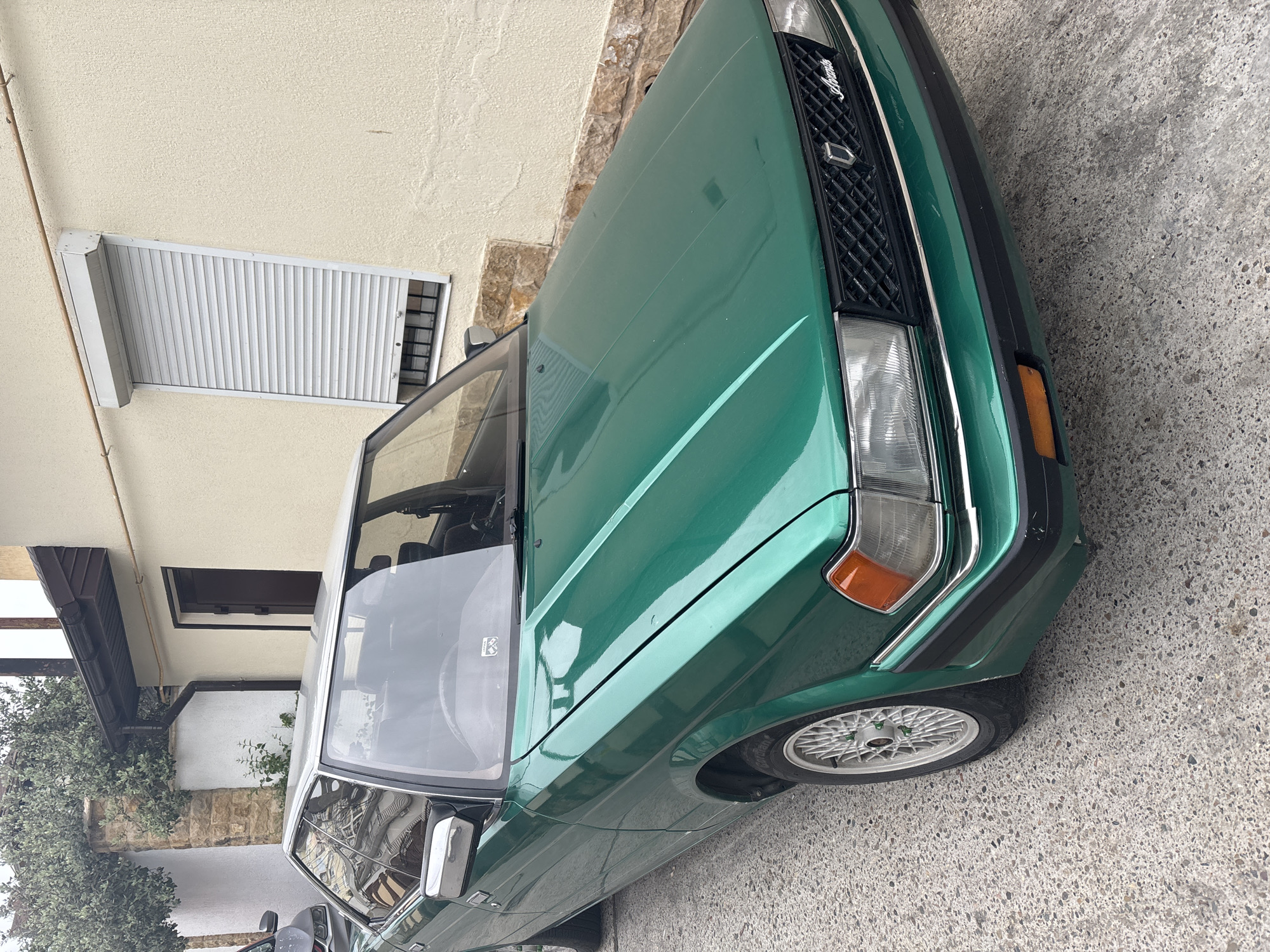 The image depicts the front corner of a green vintage car parked on a concrete surface next to a building. The car's headlight, grill, and wheel are visible, indicating it is a classic vehicle in seemingly good condition. The building features a beige stucco wall with a white shuttered window. The setting appears to be a narrow street or alley. There is no verifiable information about the location being Sochi, Russia.